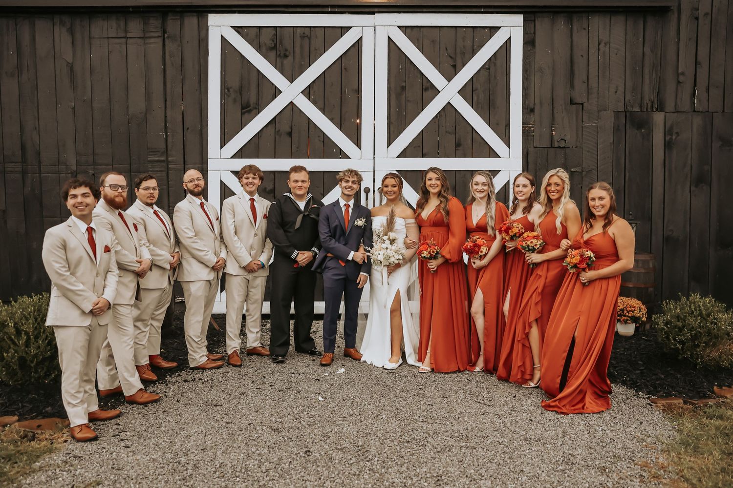 Wedding party in red dresses and tan suits poses in front of rustic barn doors.