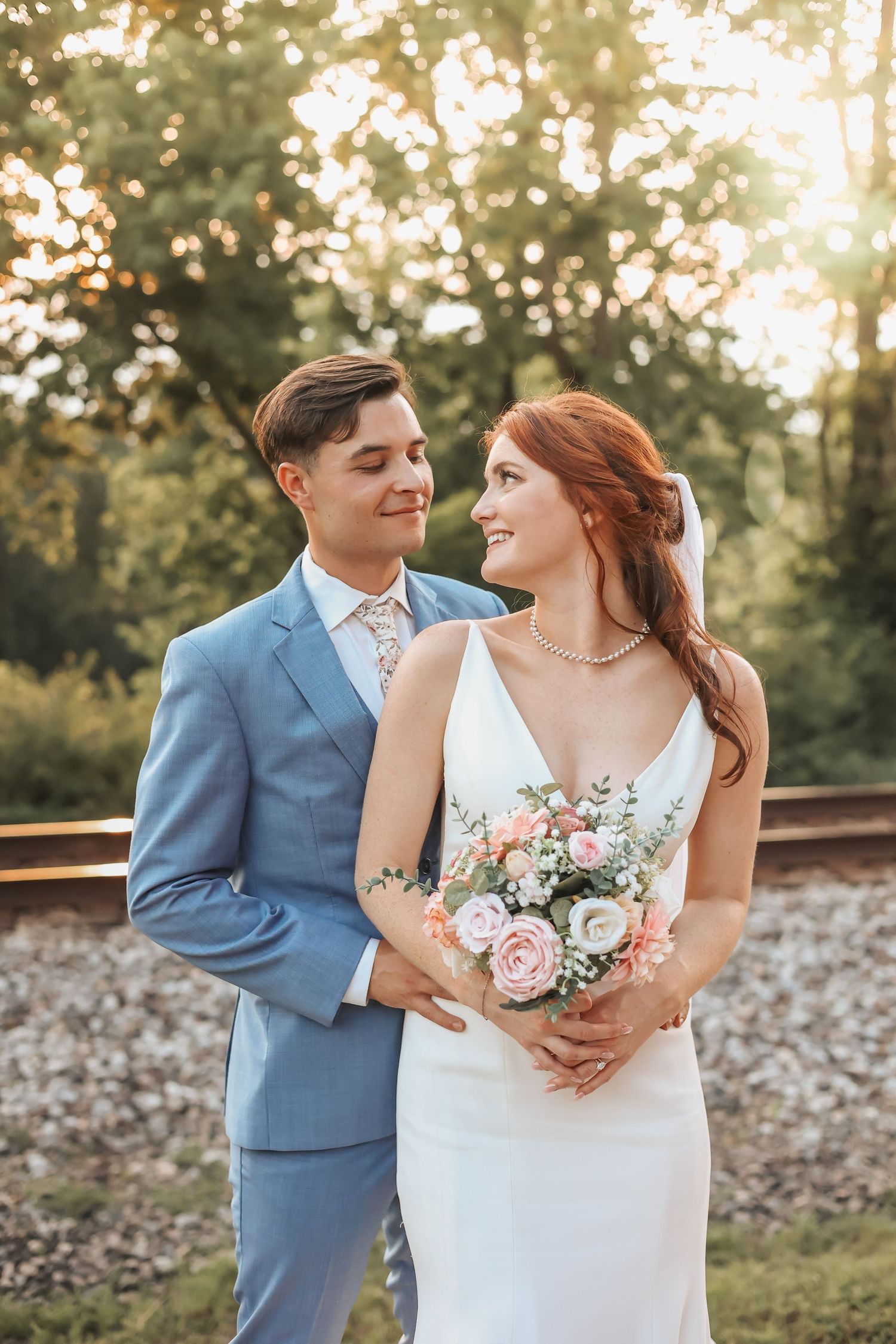 Newlyweds in elegant attire pose together on train tracks during golden hour.