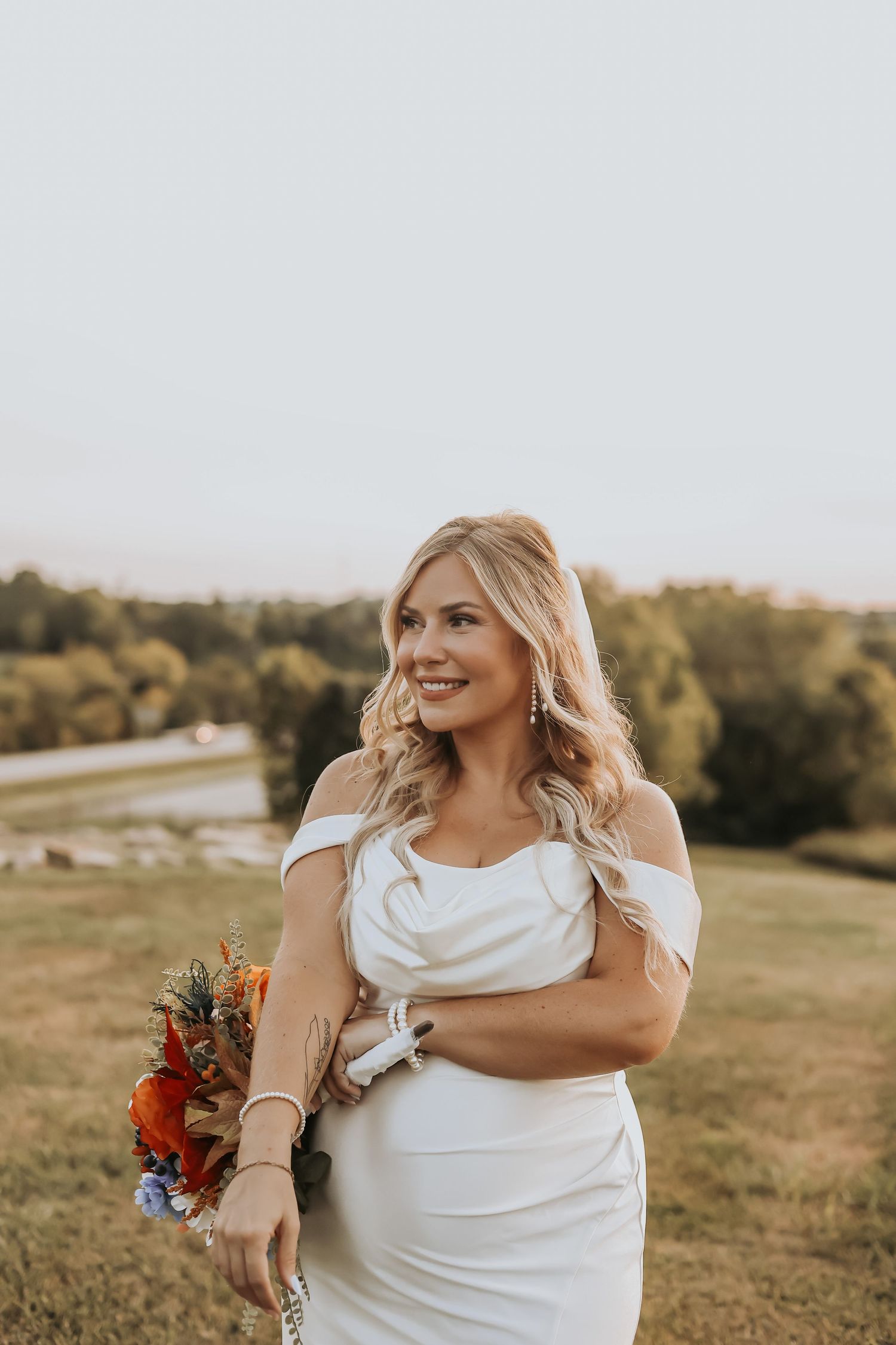 Bride in off-shoulder gown smiles while holding vibrant bouquet outdoors.