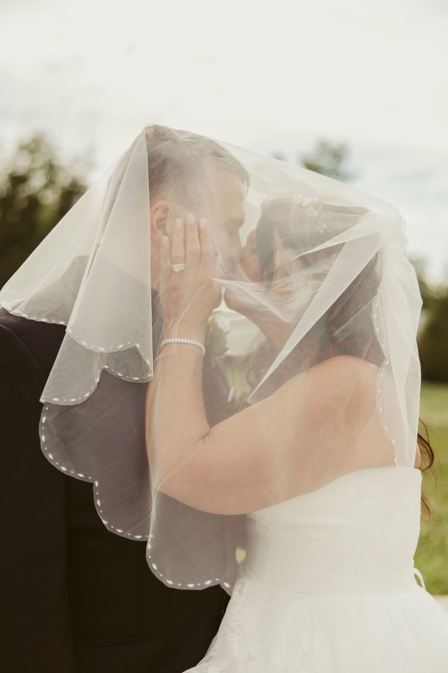 Bride and groom share a romantic kiss hidden beneath the bride's flowing white veil outdoors.