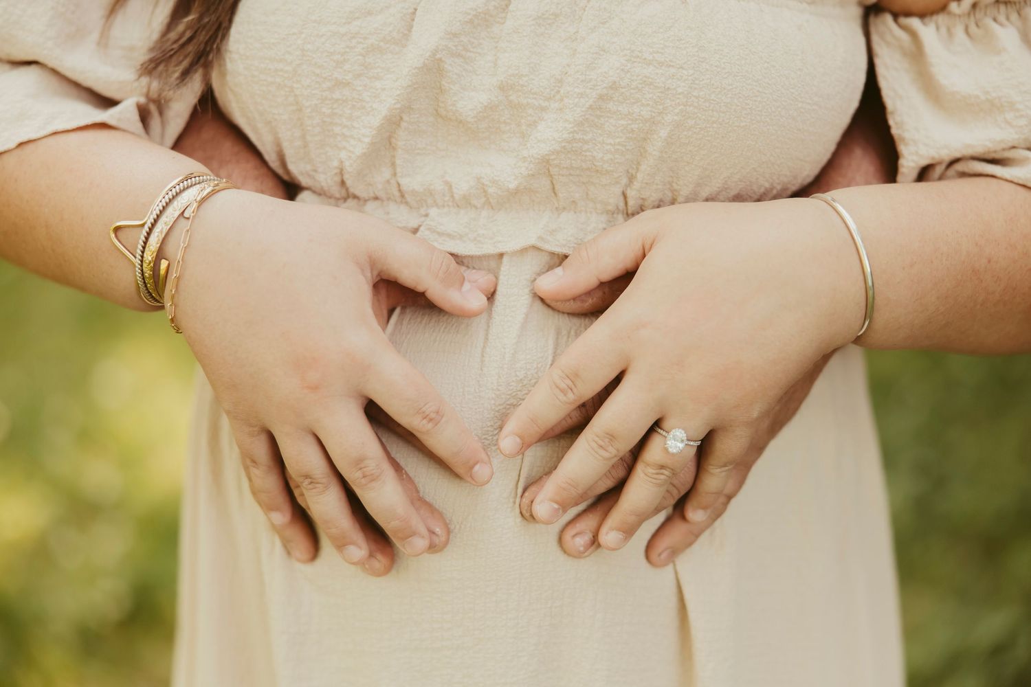 Couple forming heart shape with hands on pregnant woman's belly, wearing gold bracelets and diamond ring.