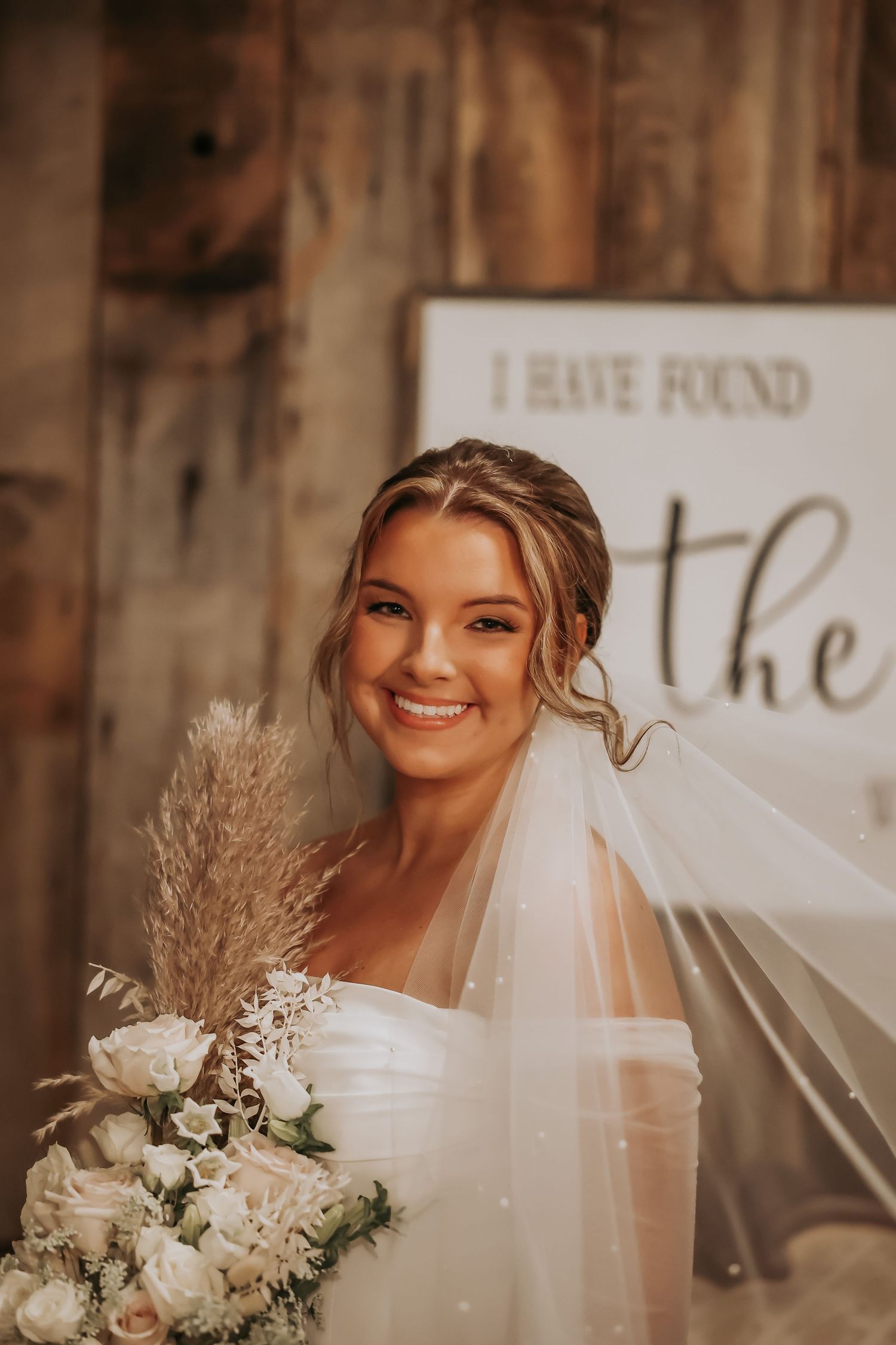 Bridal portrait against rustic wall with 'The One' sign featuring pampas grass bouquet.