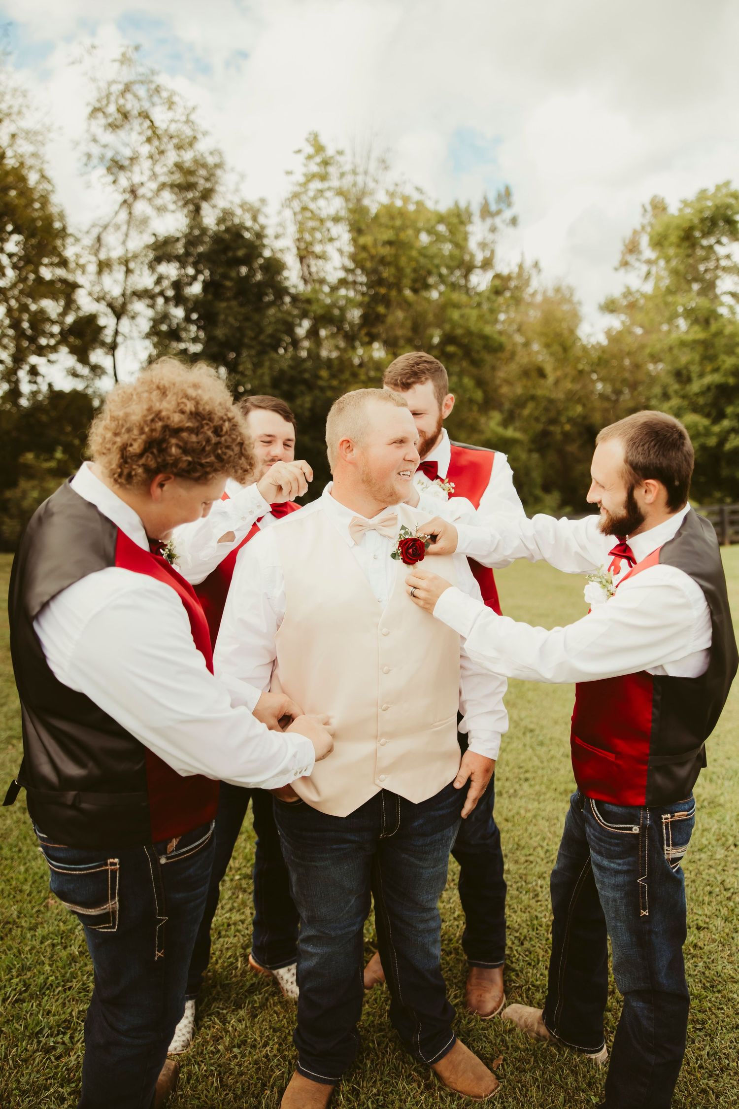 Groom in beige vest and jeans surrounded by groomsmen in red vests on green lawn.