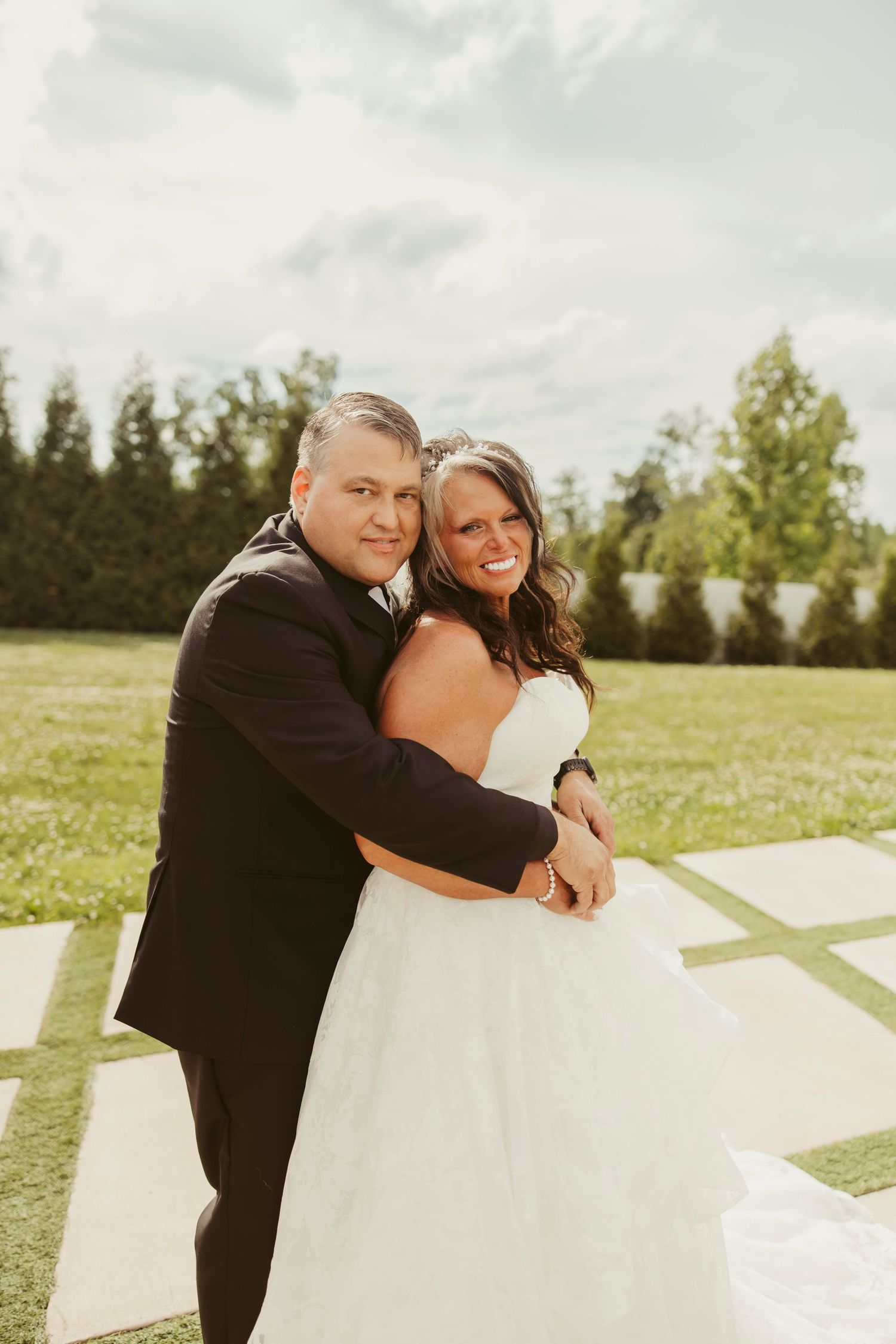 Bride and groom embracing and smiling outdoors on a sunny day, with lush green trees in the background.