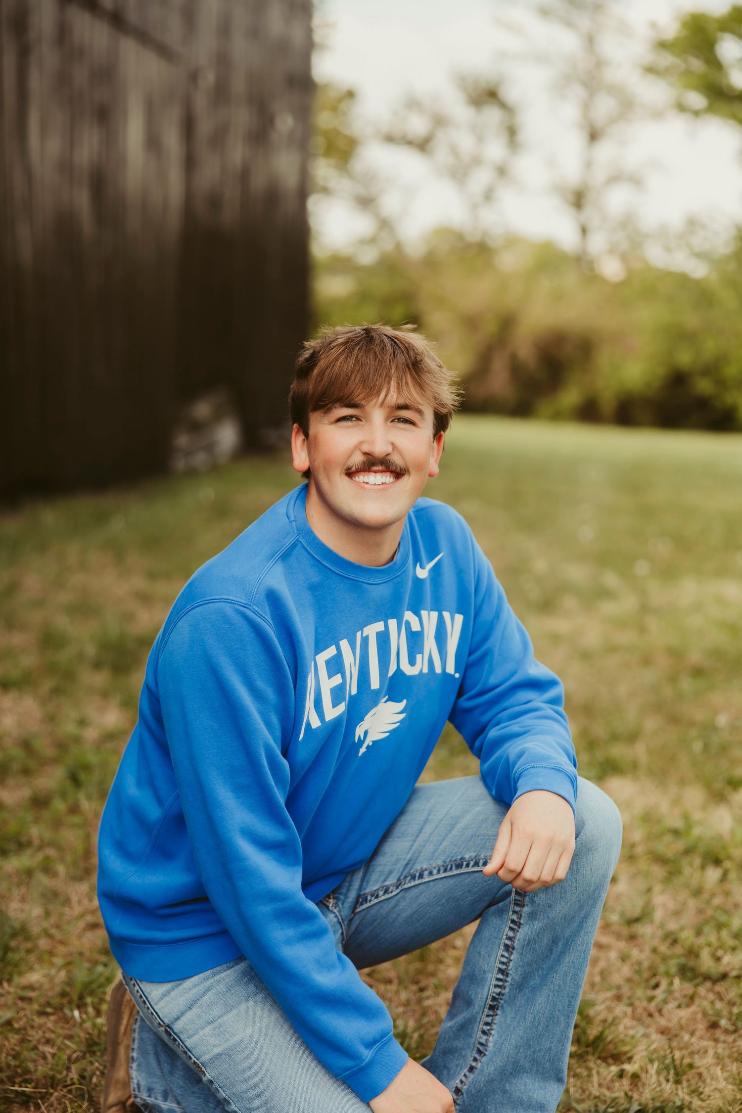 Young man smiling in blue Kentucky Nike sweatshirt and jeans, sitting outdoors near a barn.