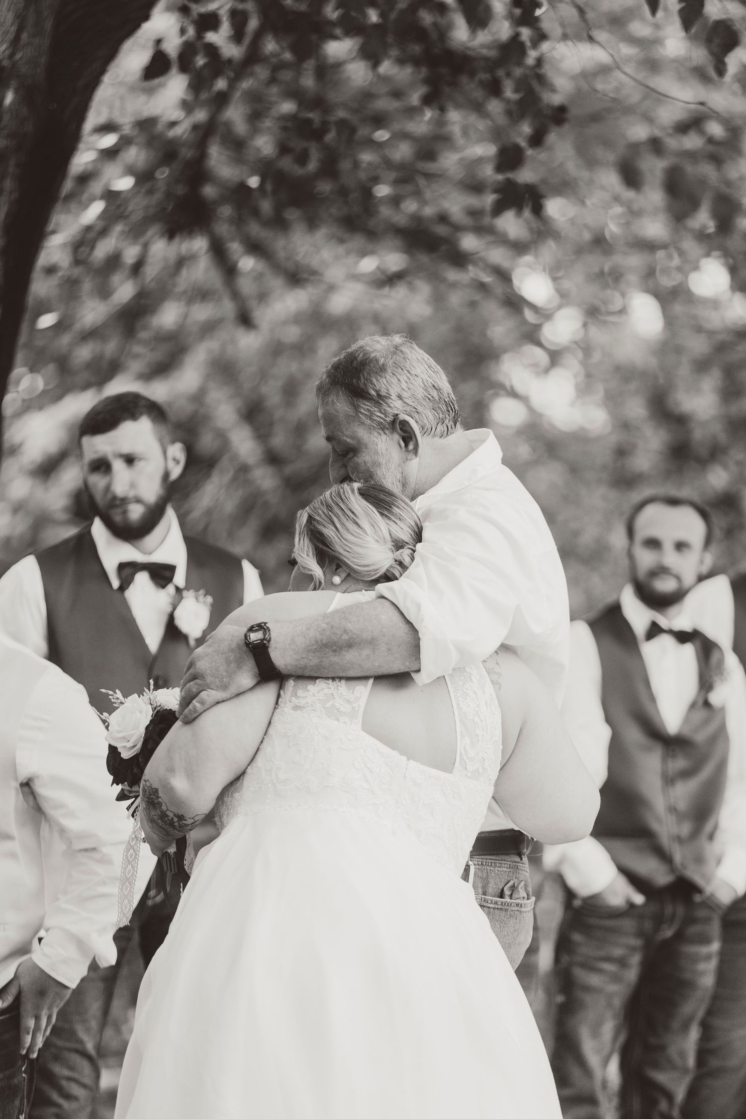 Black and white photo of bride and groom sharing emotional hug during outdoor wedding ceremony with groomsmen in background.