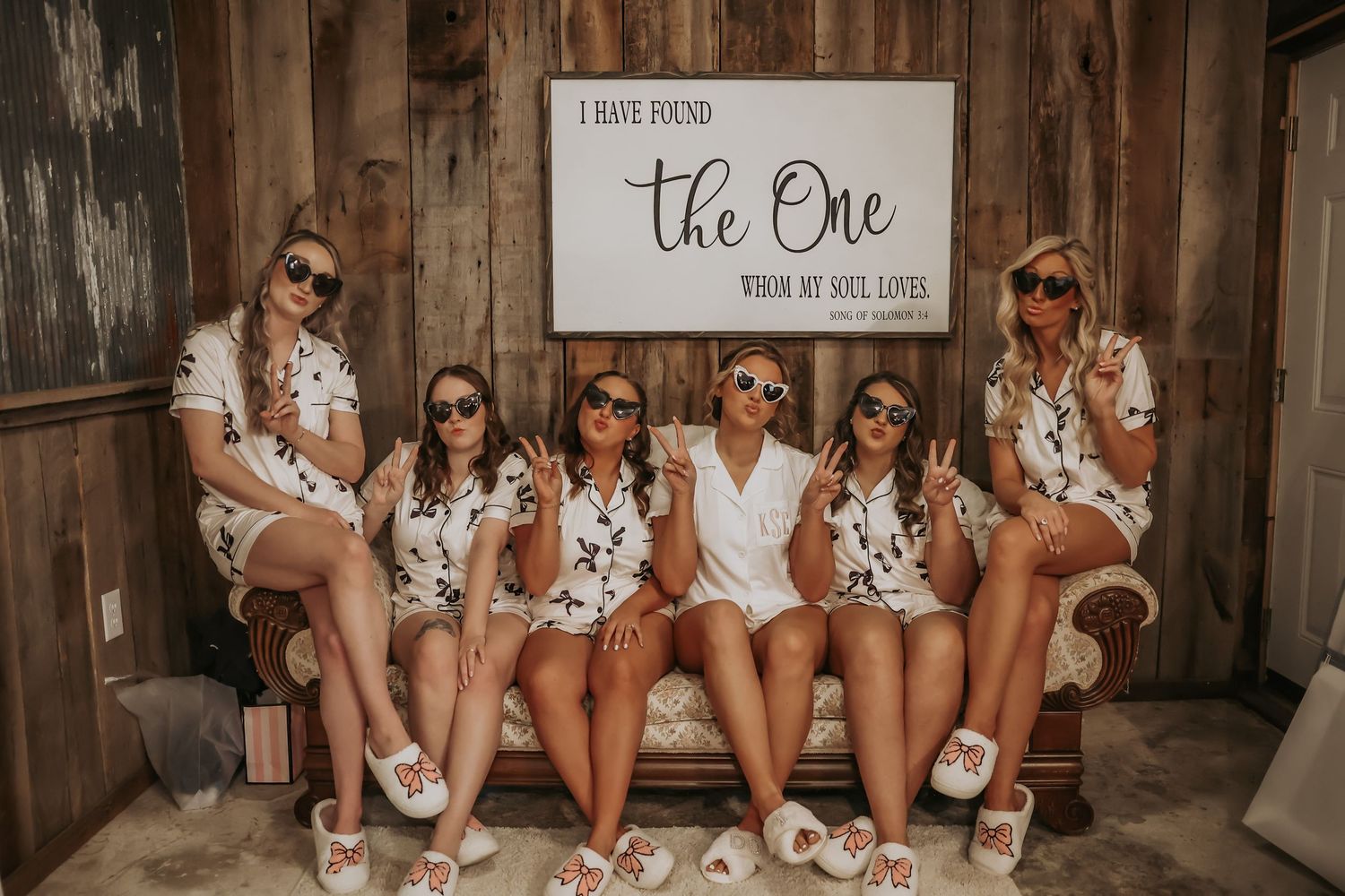 Wedding party in matching outfits poses with 'The One' sign in rustic venue.
