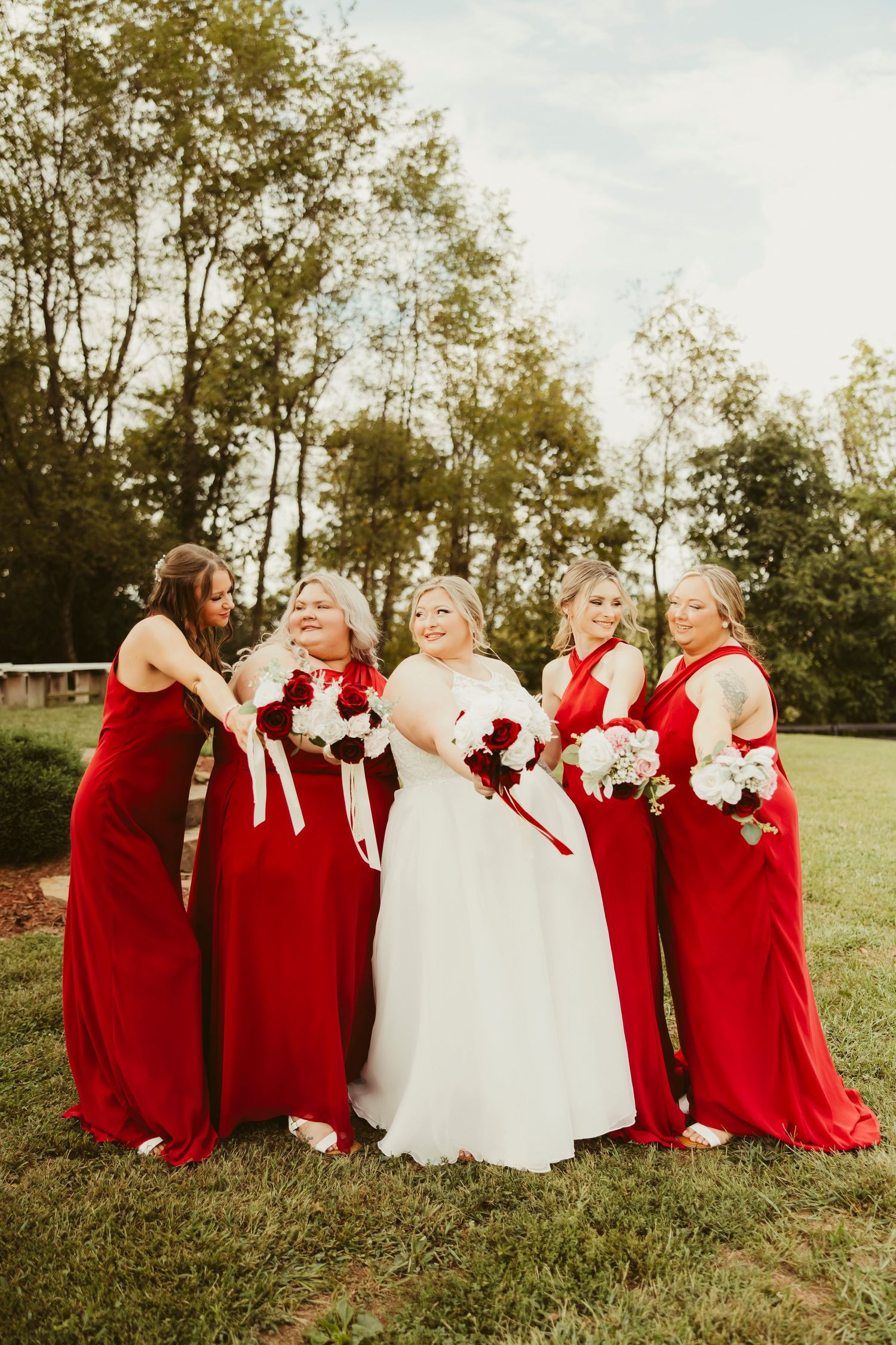 Bride in white gown poses with four bridesmaids in red dresses holding bouquets outdoors.