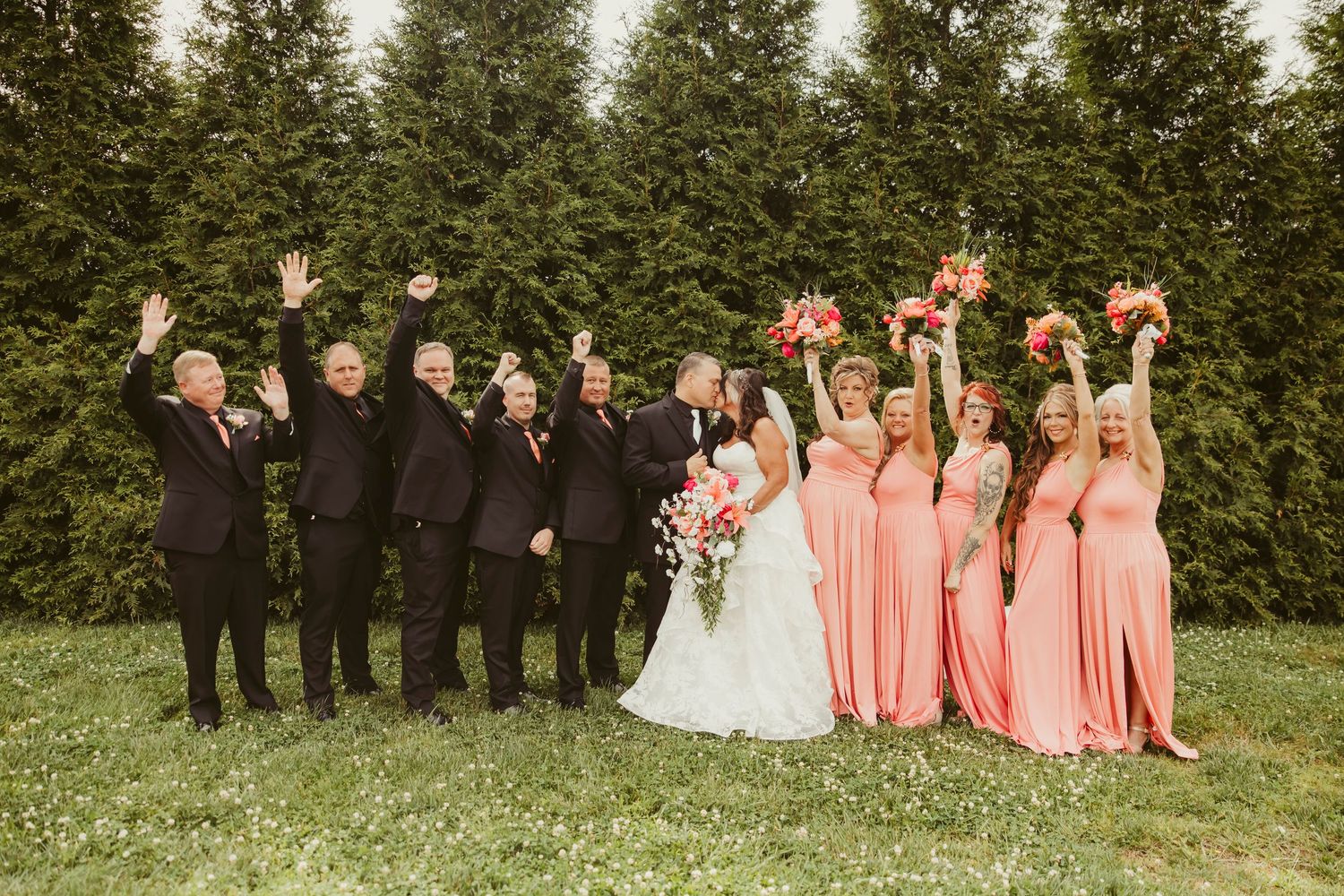 Wedding party in coral dresses and black suits celebrating outdoors with raised hands before evergreen trees.