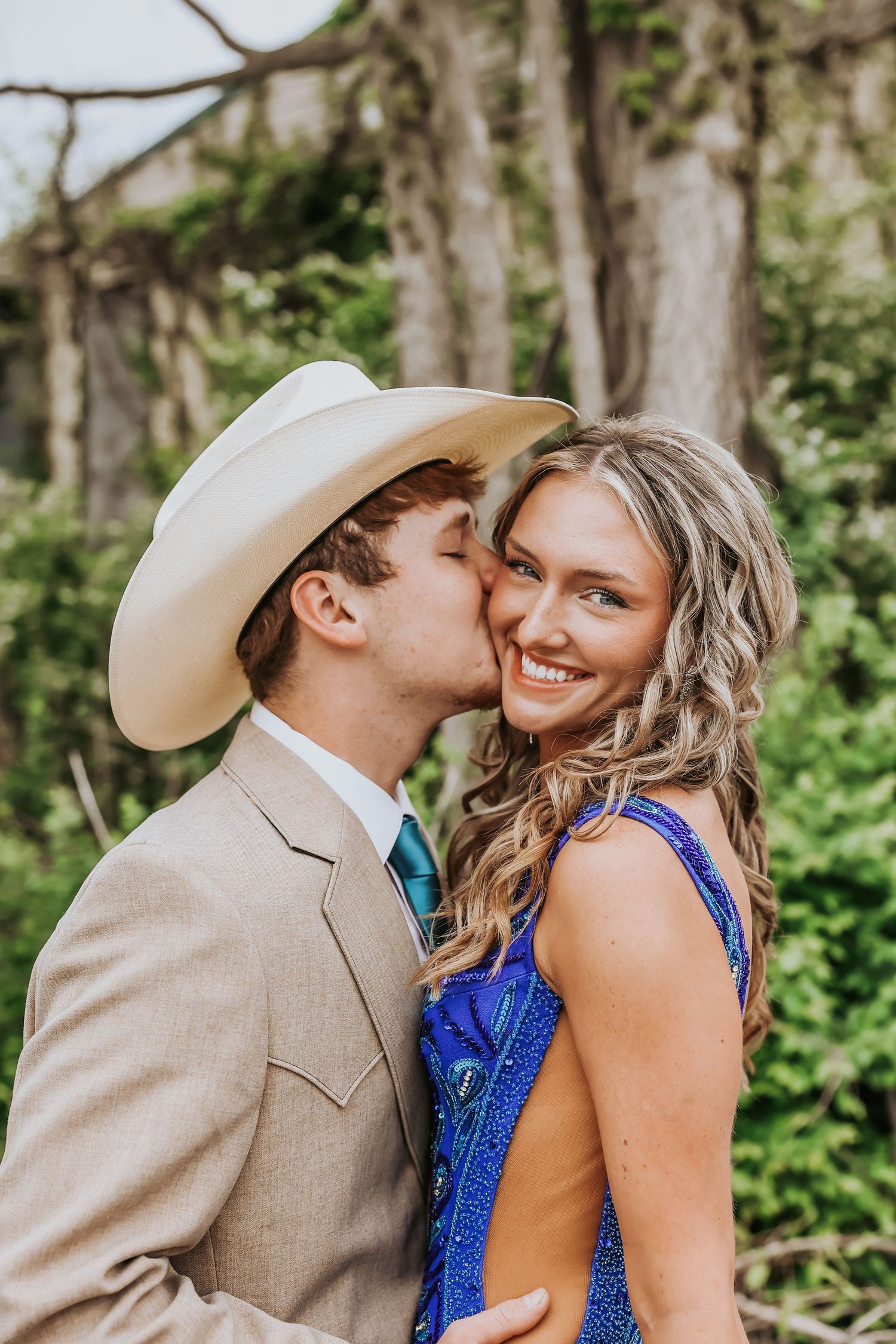 A couple in formal attire share an intimate moment outdoors, with him in a tan suit and cowboy hat and her in a blue dress.