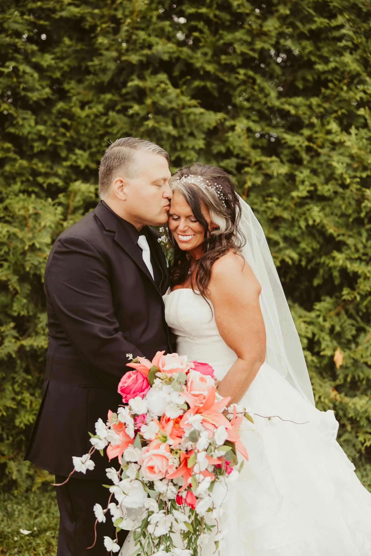 Groom in black suit kissing bride on cheek as she smiles holding colorful pink coral wedding bouquet outdoors.