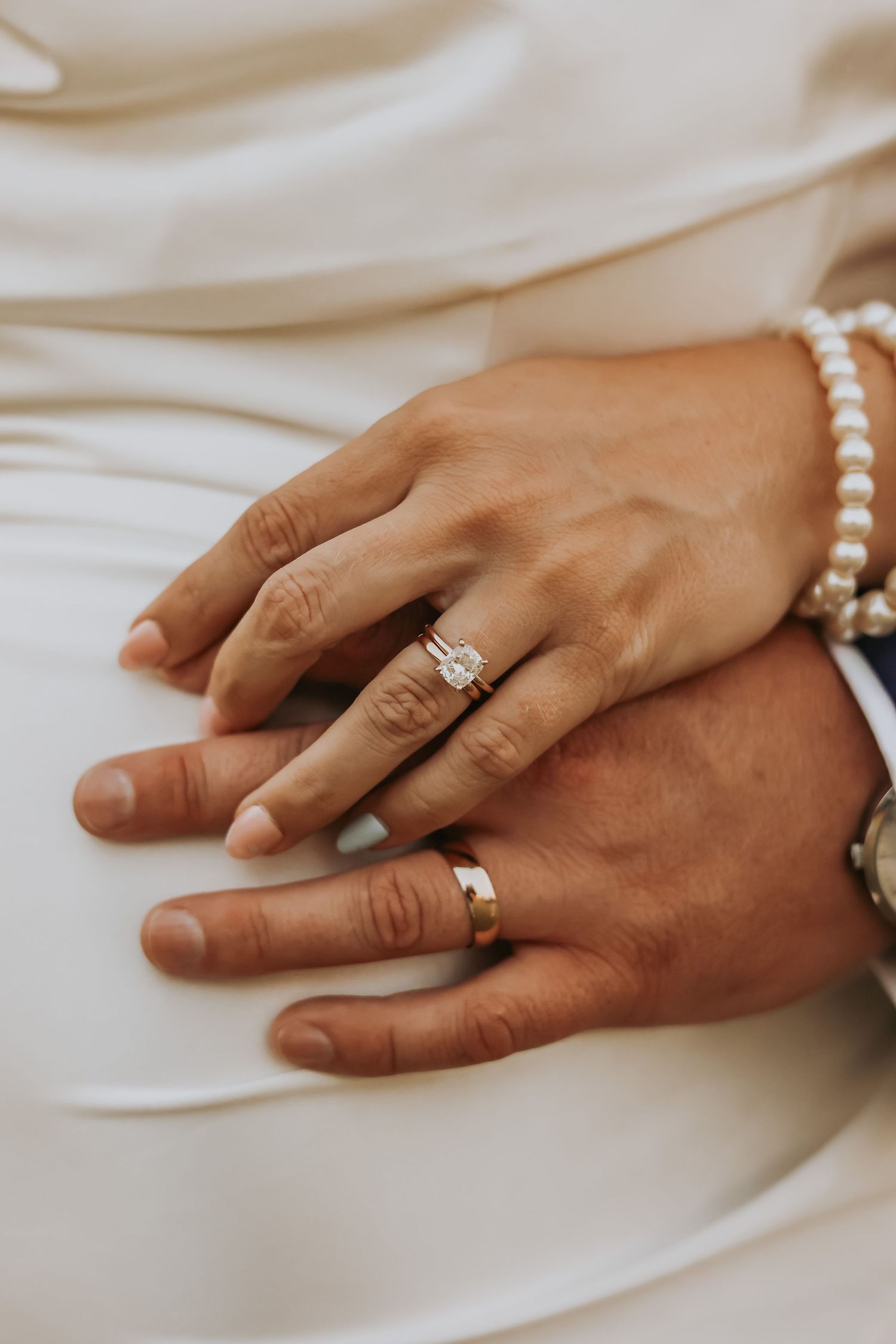 Close-up of newlywed hands with wedding rings and pearl bracelet on white fabric.