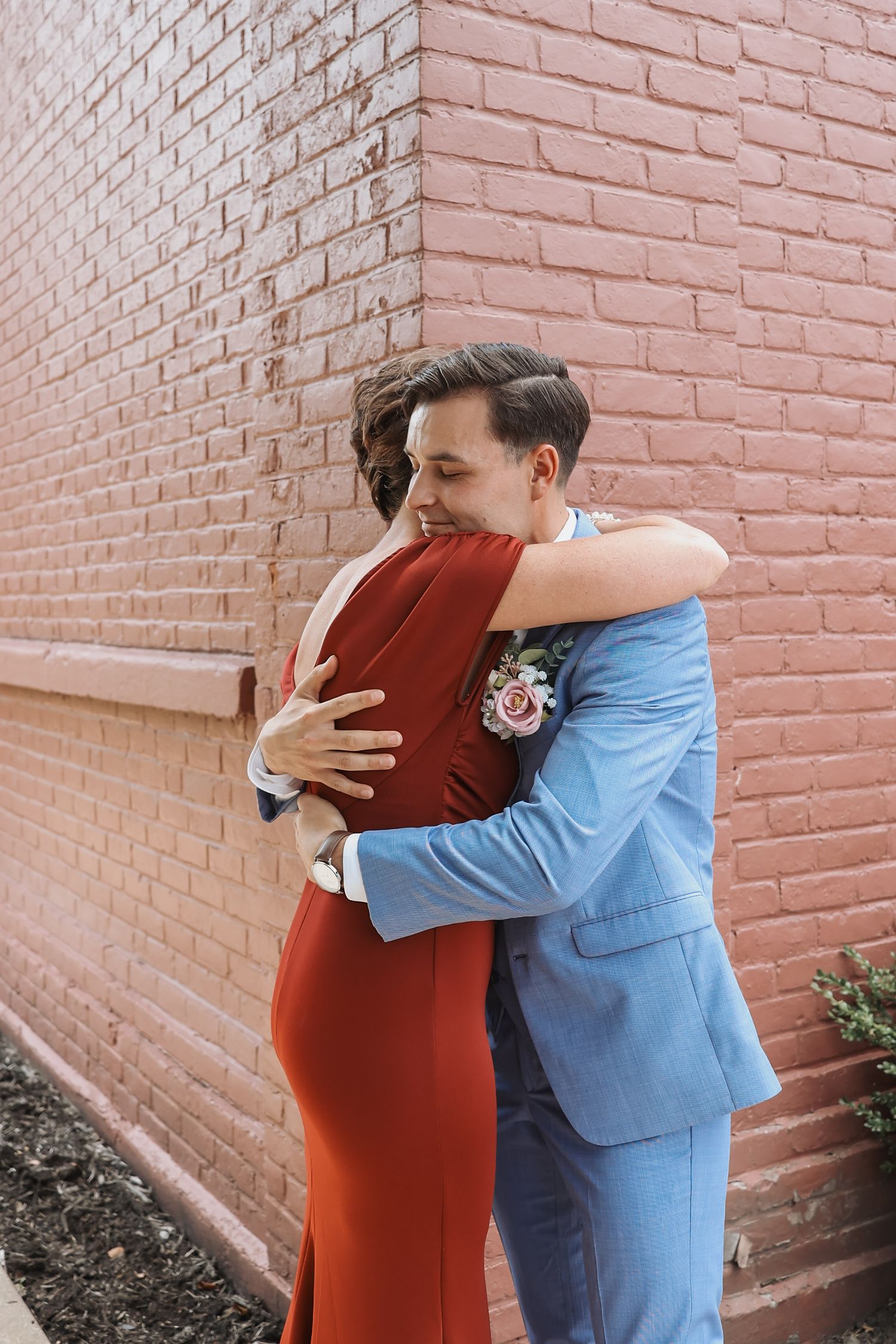A couple embraces in front of a pink brick wall, with one wearing a red dress and the other in a blue suit.