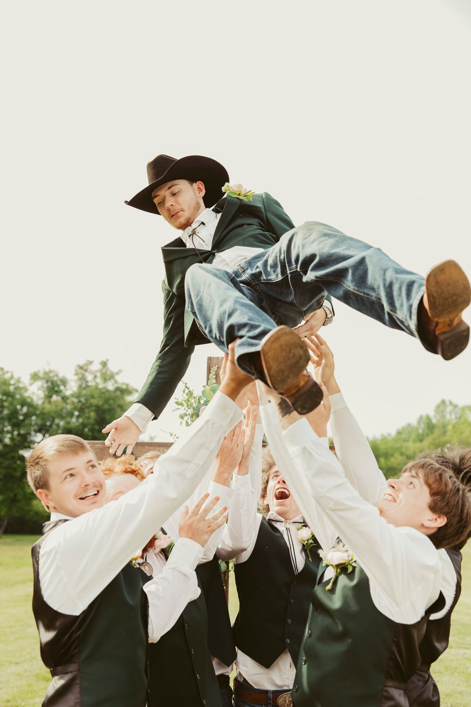 Groomsmen tossing the groom in the air outdoors at a wedding, with the groom wearing a cowboy hat and boots.