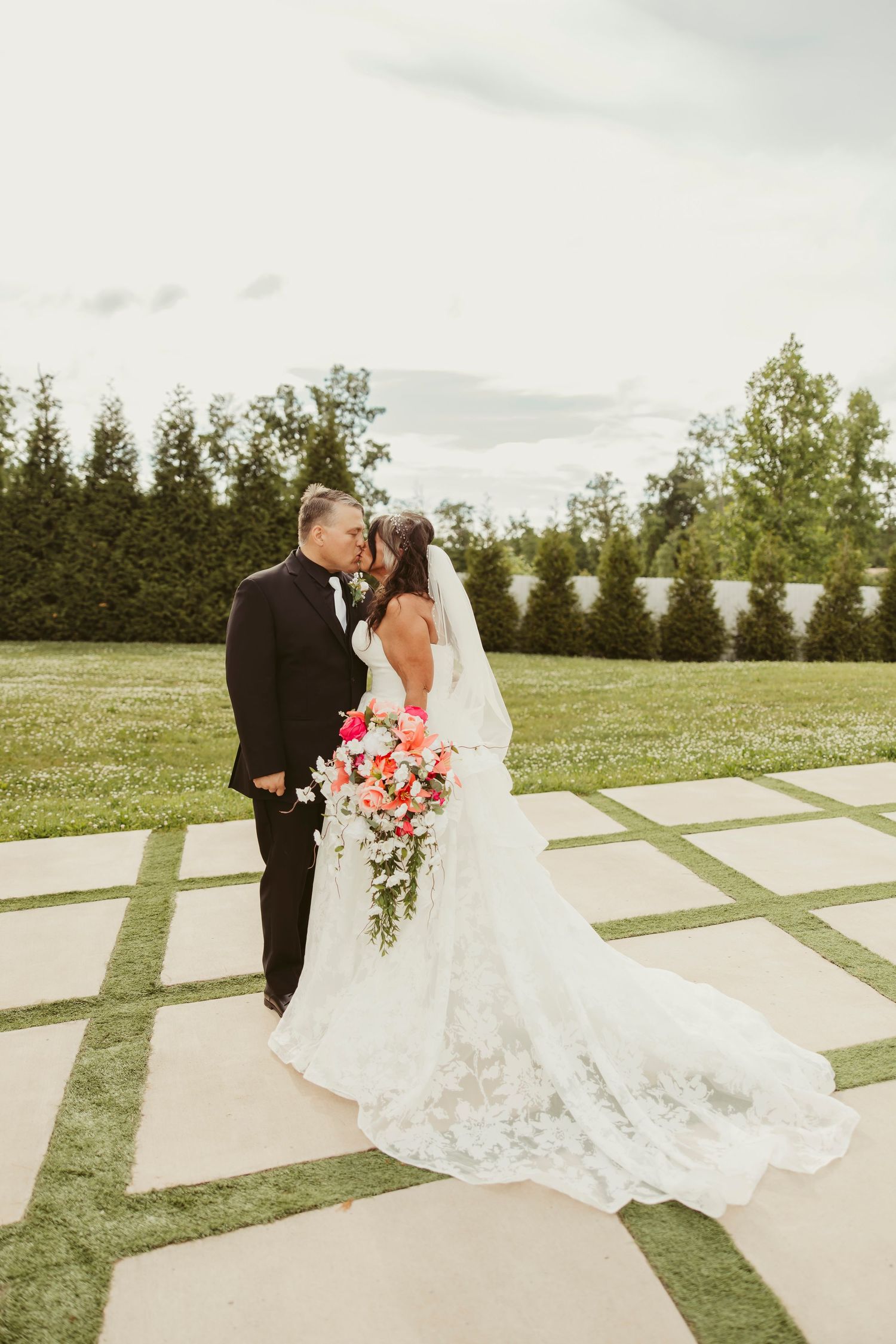 Bride and groom share a kiss on a stone patio, her lace gown and veil flowing, holding a vibrant coral bouquet.