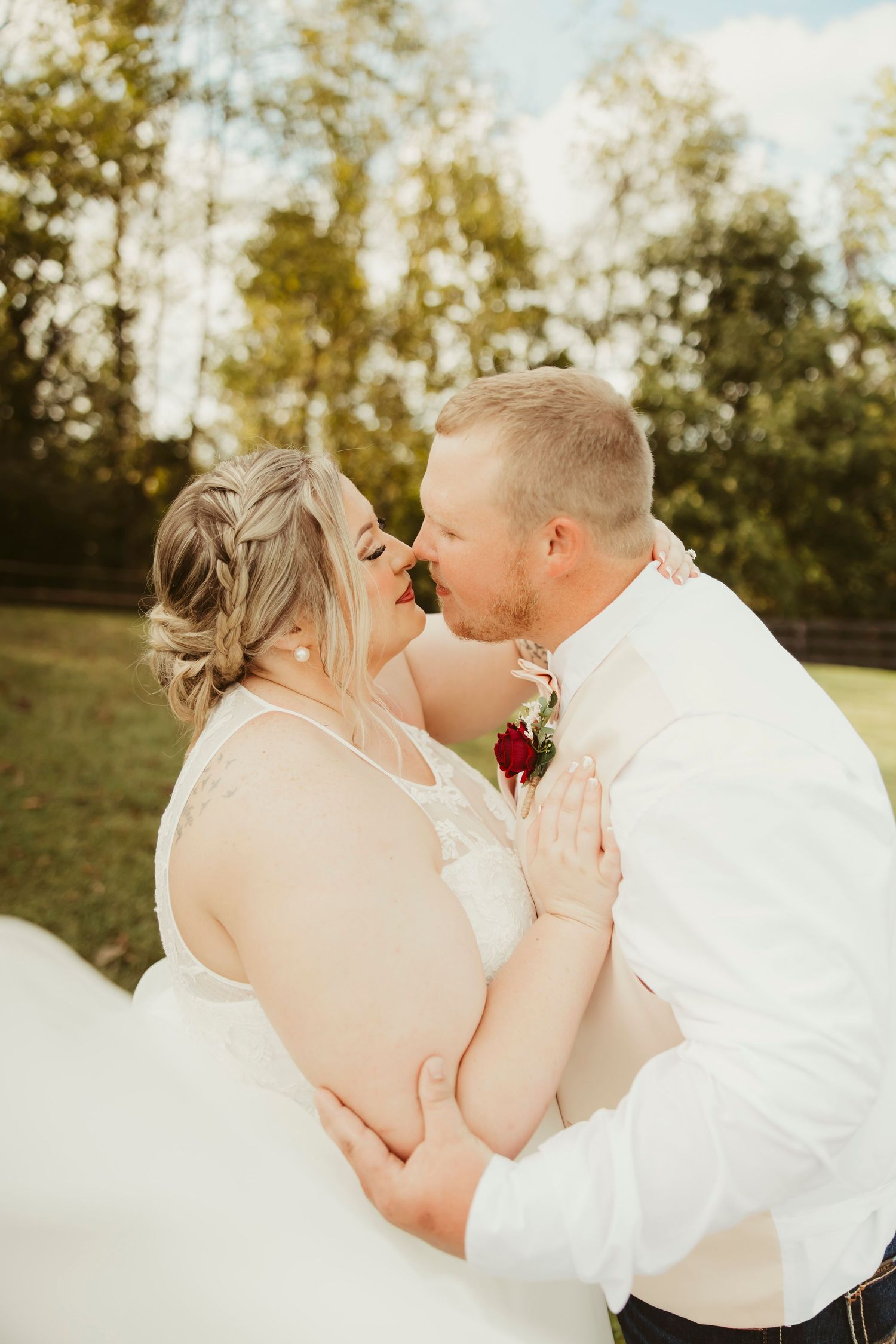 Bride and groom share romantic almost-kiss embrace outdoors, groom wearing red rose boutonniere on white shirt.