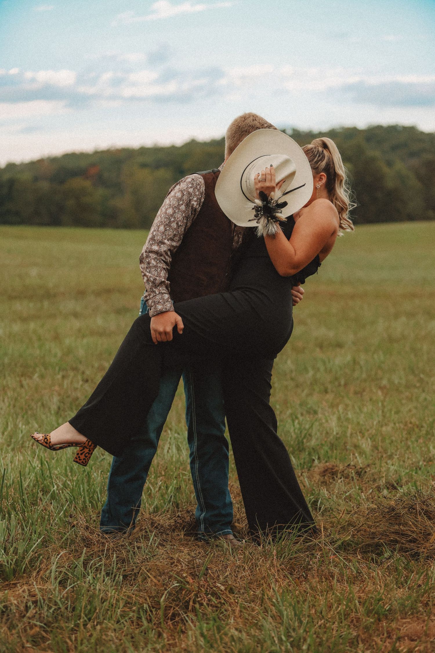 A romantic couple embraces in a grassy field at sunset with one person wearing a cowboy hat and a floral dress.