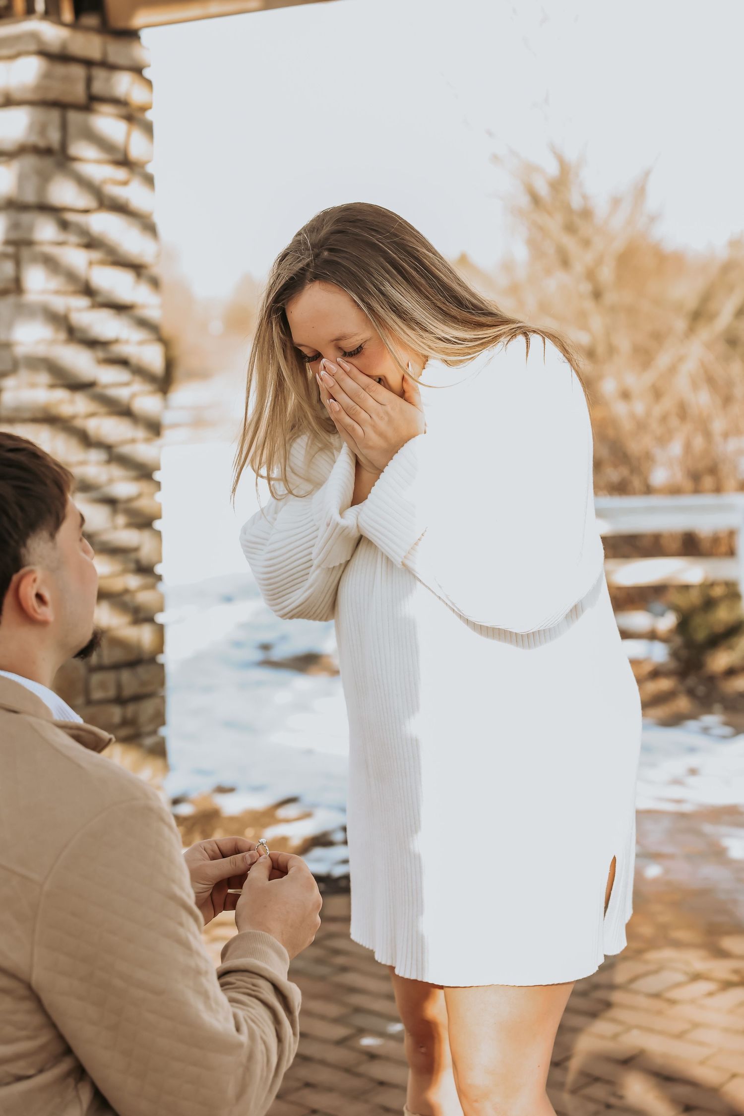A surprise proposal moment captured outdoors in winter with one person kneeling while holding a ring in front of another in white.