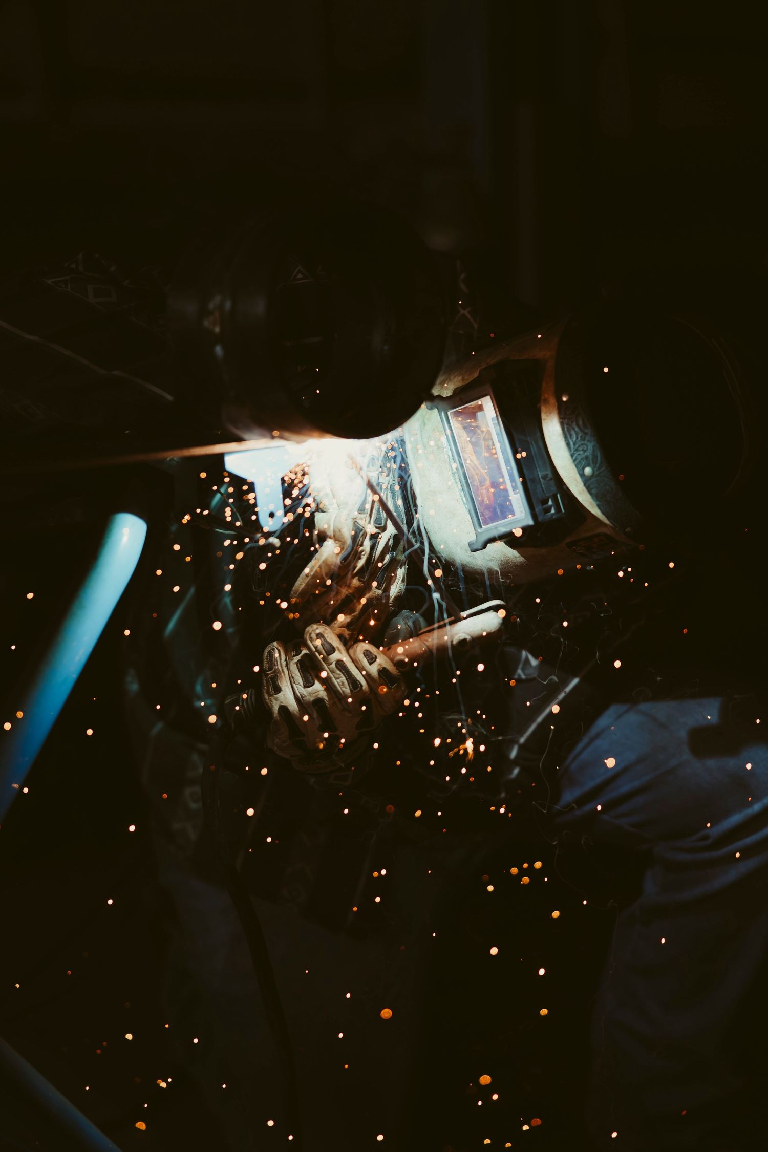 A welder in a protective helmet works with bright sparks flying in a dark industrial workshop.