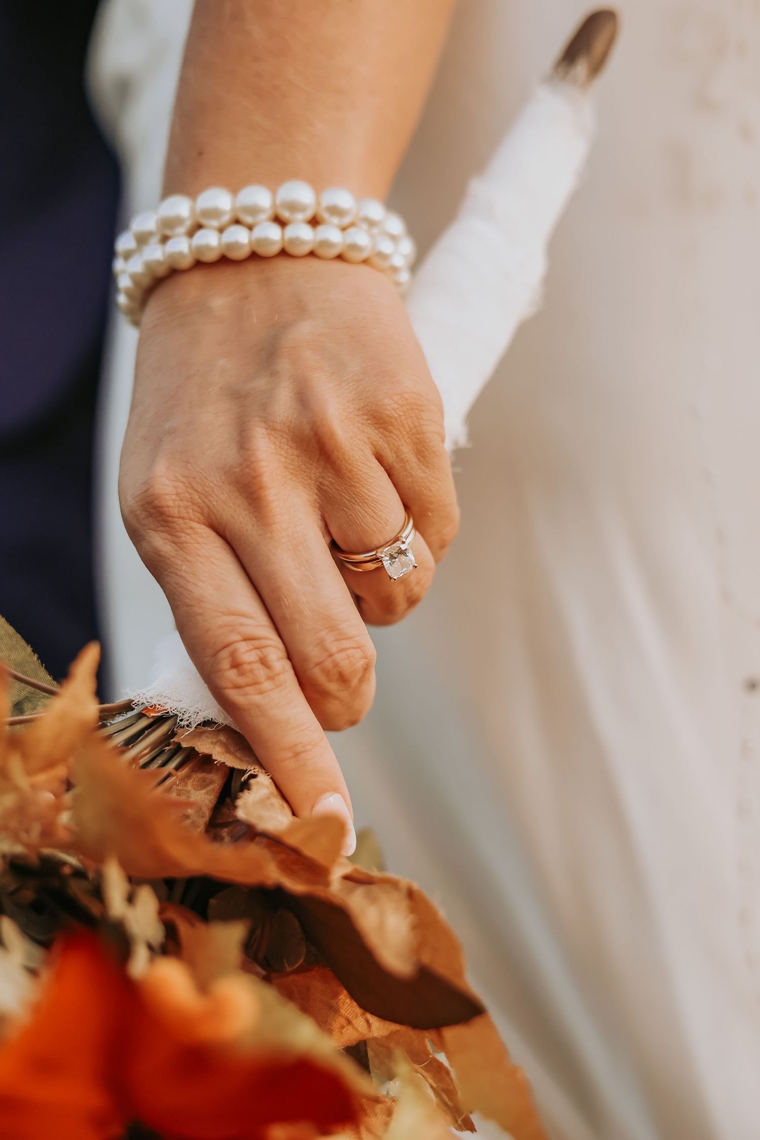 Detail shot of bride's hand with engagement ring, pearl bracelet, and autumn bouquet.