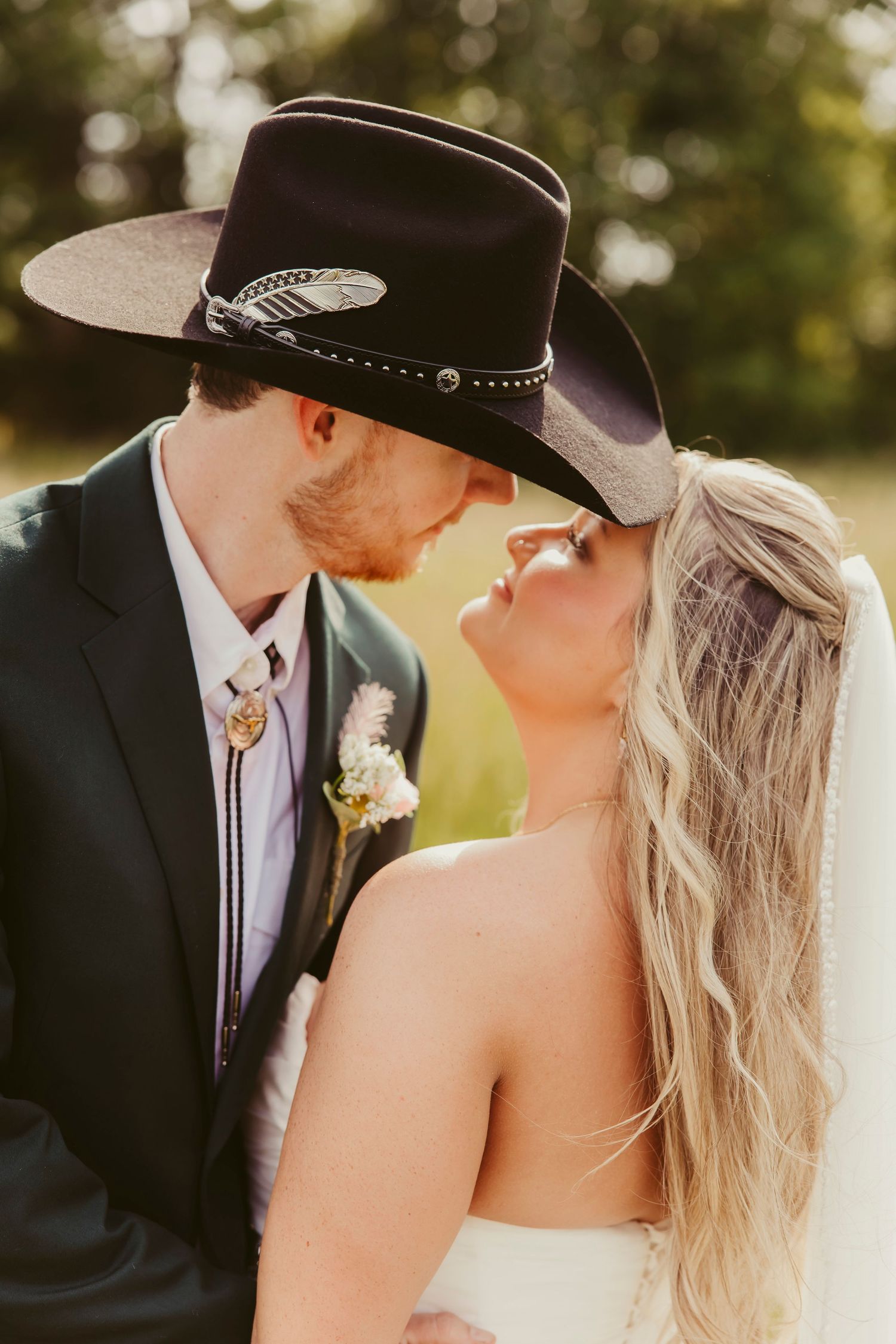 A bride and groom share a romantic kiss under a black cowboy hat at their rustic outdoor wedding ceremony.