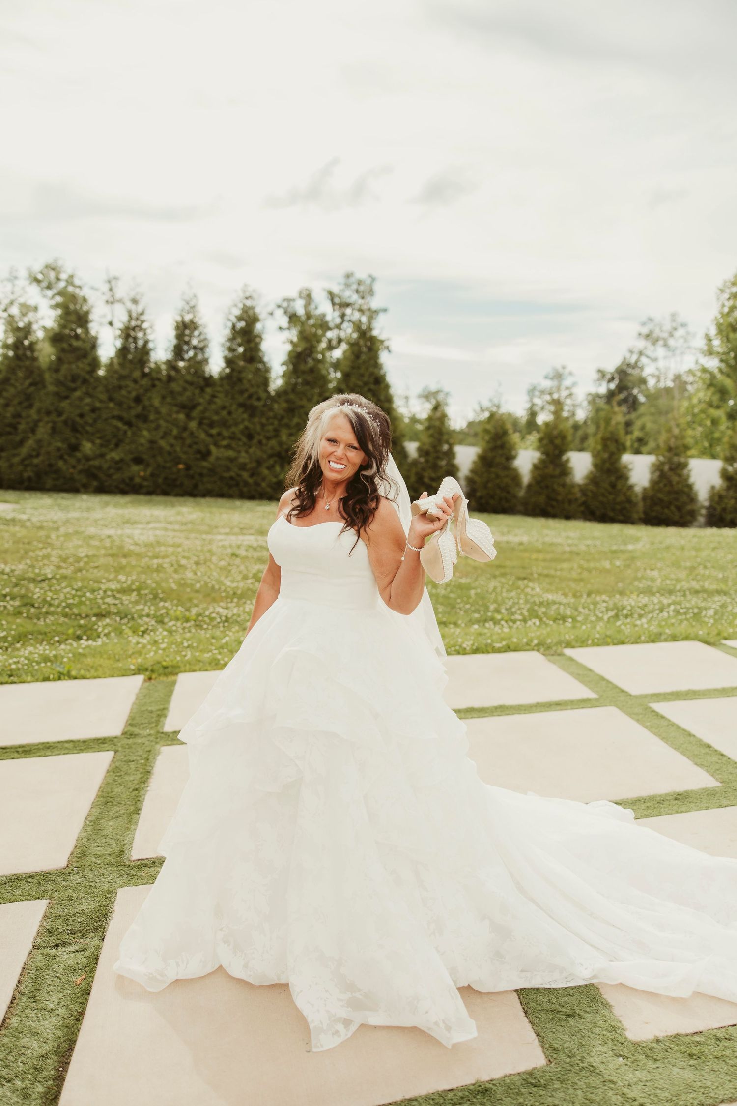 Bride in a full ball gown wedding dress twirling outdoors on a sunny day, holding her shoes and smiling.