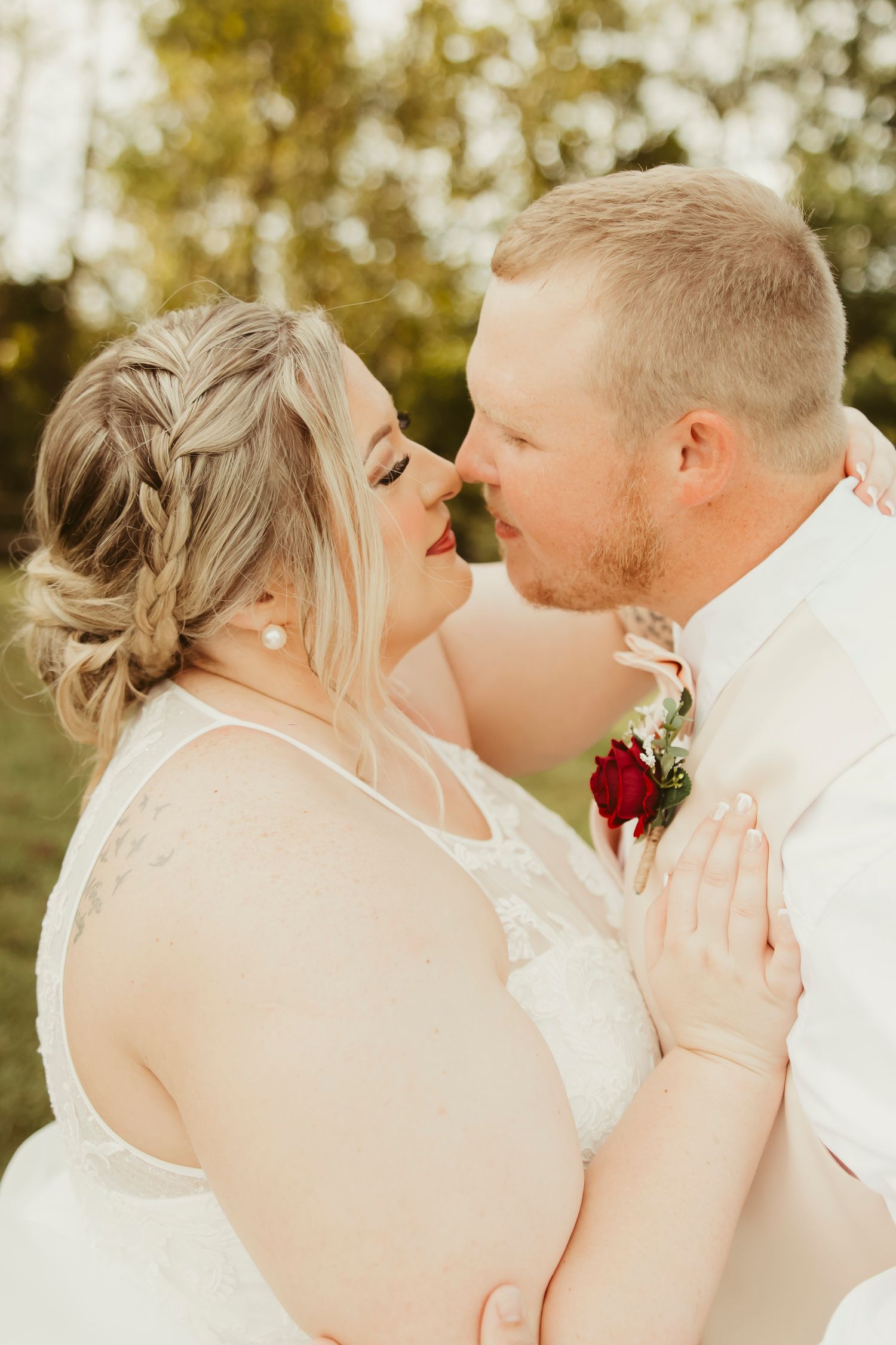 Bride and groom share a romantic nose-to-nose moment, bride in lace gown with braided updo, groom in white suit with red rose boutonniere.