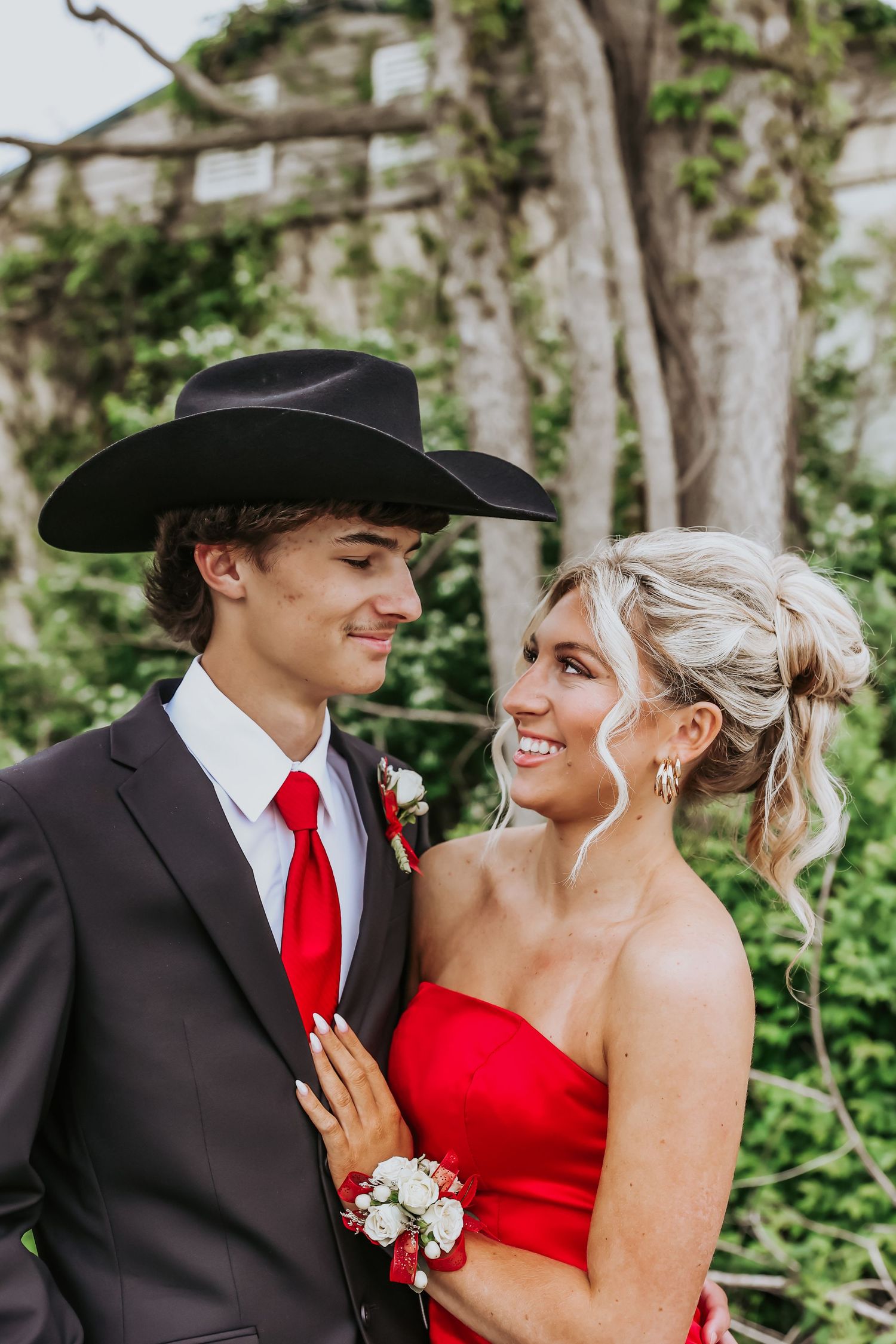 A couple dressed in formal prom attire with a black cowboy hat and red dress smile at each other outdoors.