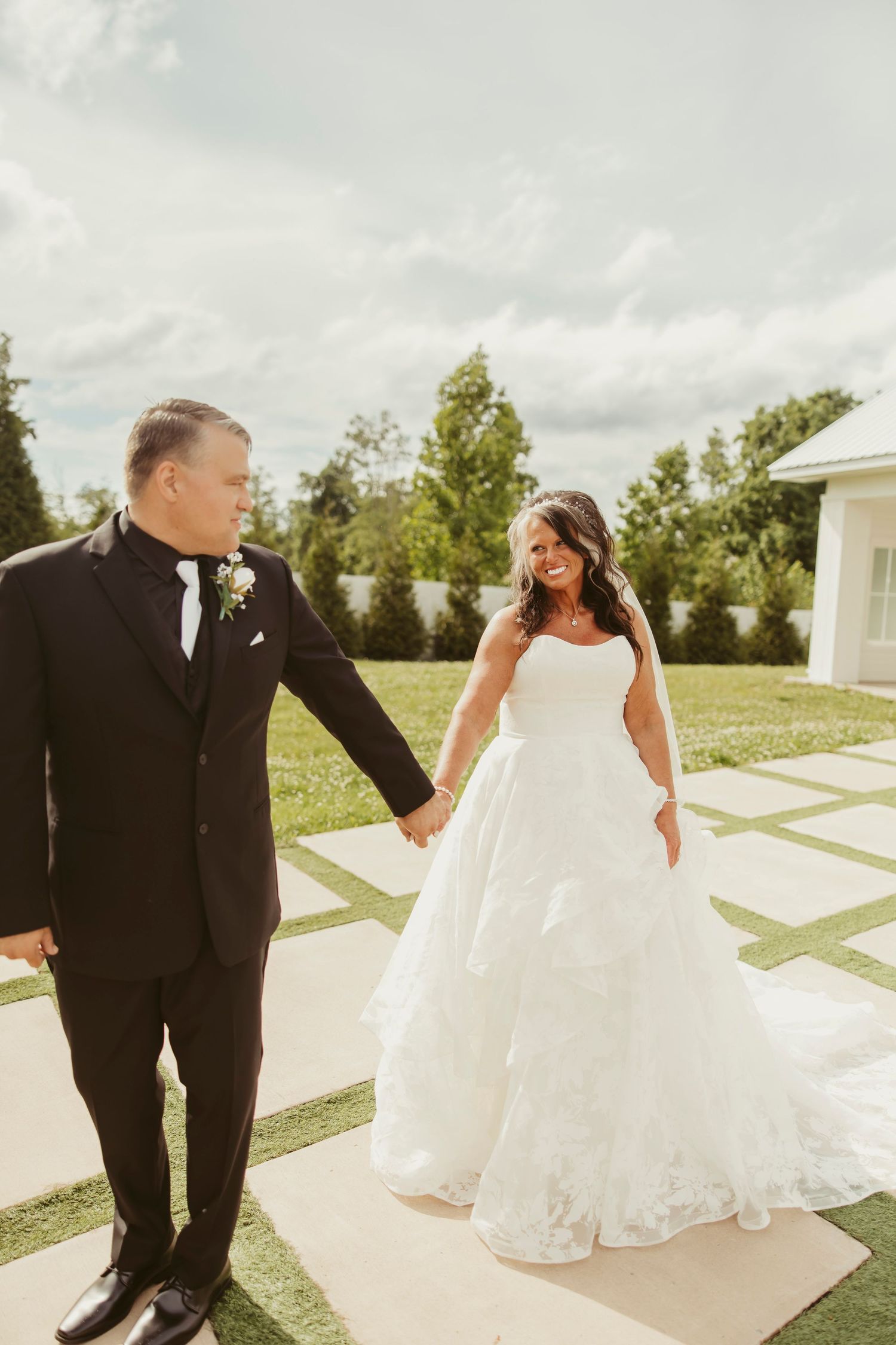 Bride in white ball gown and groom in black tuxedo hold hands and smile at each other outdoors on their wedding day.