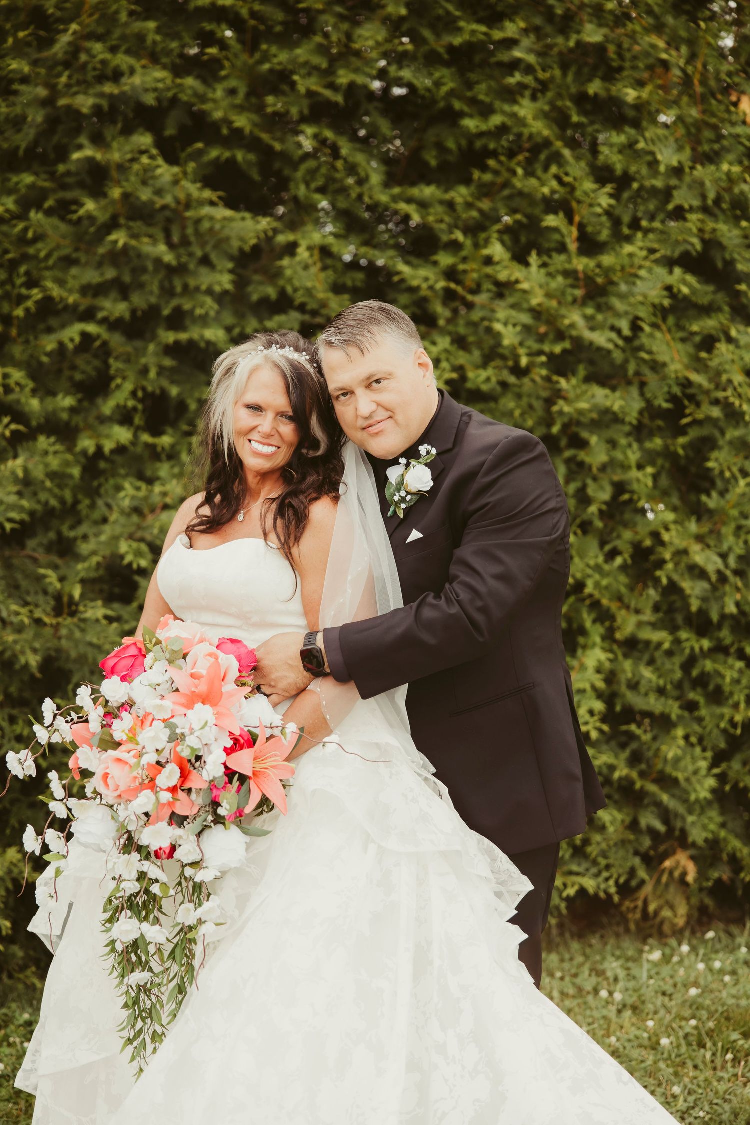 Bride and groom pose together outdoors, she holds a coral and white bouquet, he wears a black tuxedo with boutonniere.