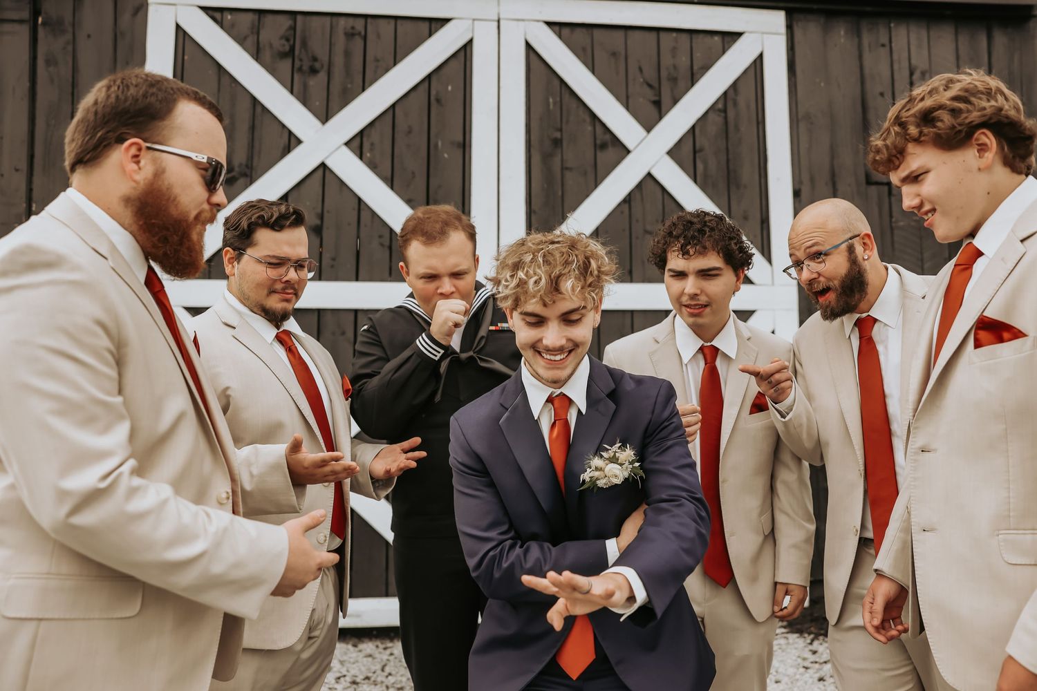 Groomsmen in light and dark suits with red ties gathered around at a rustic venue.
