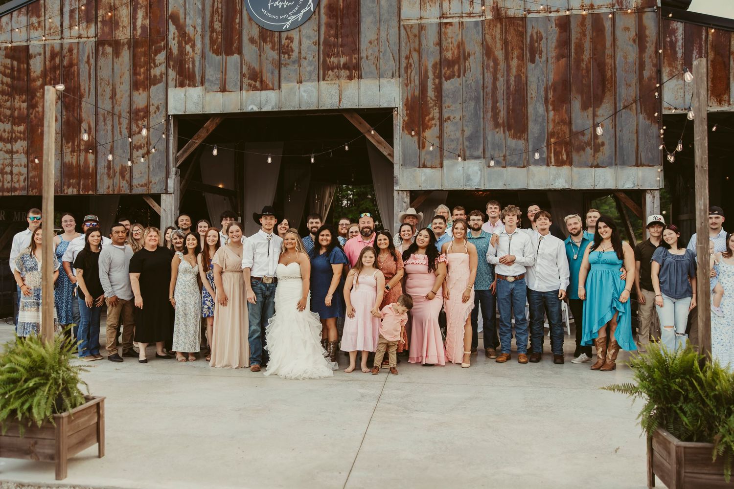 Large wedding party posing in front of a rustic barn venue with string lights and wooden doors.