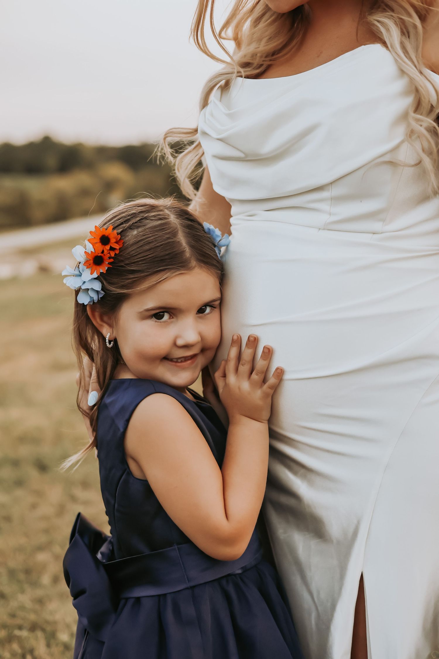 Young girl in a navy dress with orange flowers in her hair hugs a white wedding gown.