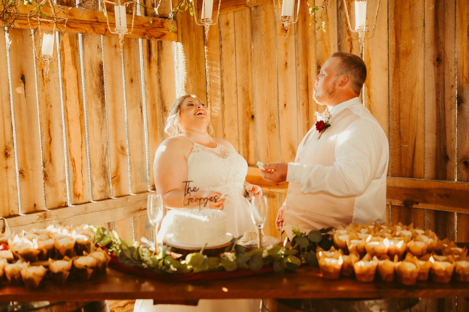 Bride and groom laugh together while cutting wedding cake at rustic barn reception venue.
