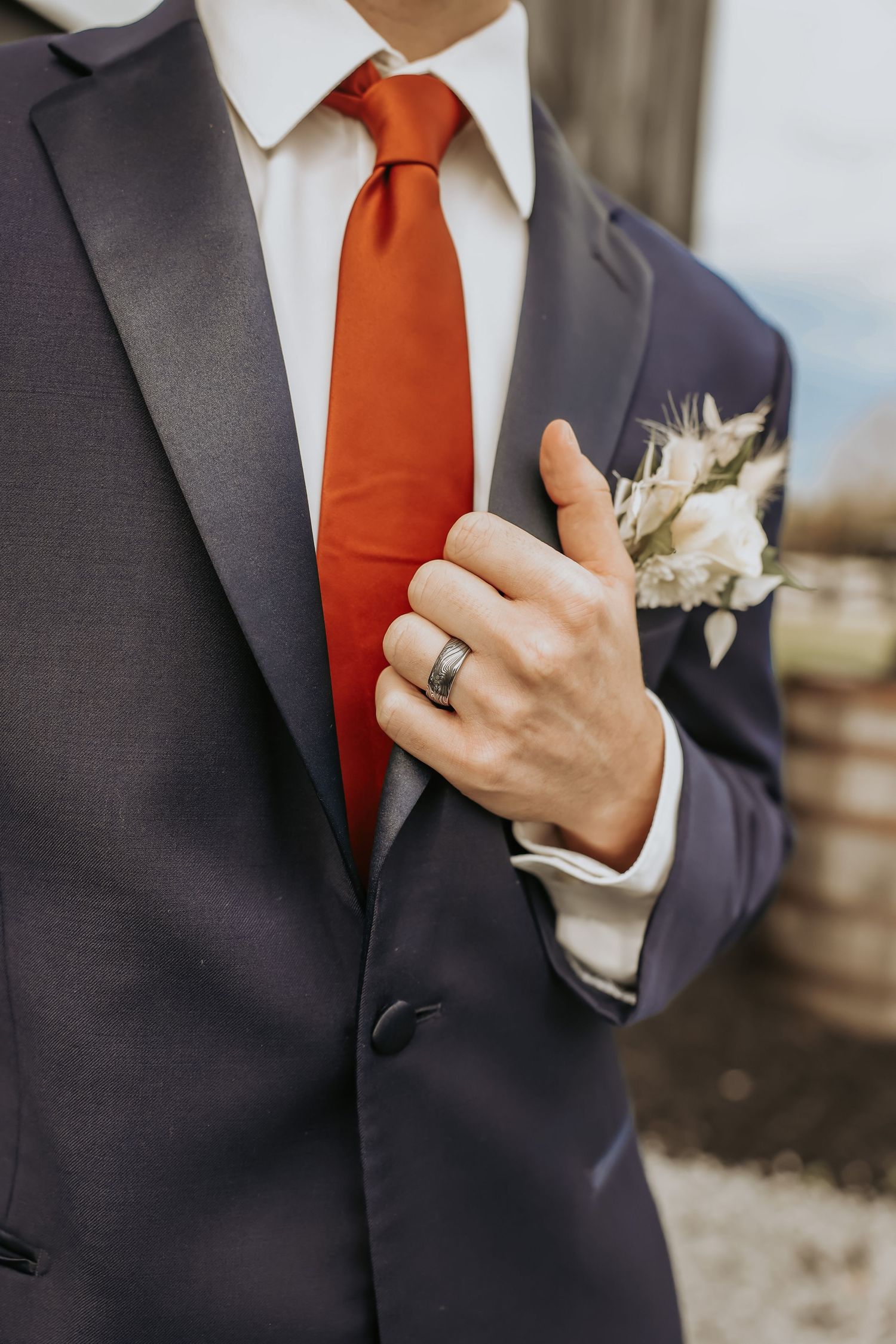 Groom's formal attire featuring charcoal suit, bright orange tie, and boutonniere with wedding ring.