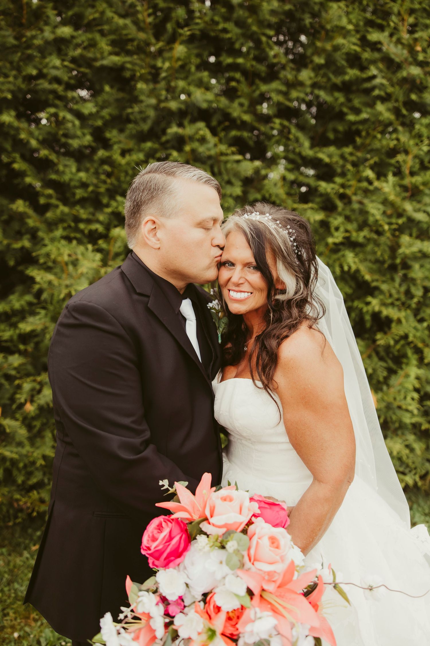 Groom in black suit kisses bride wearing white gown and veil, holding pink and coral bouquet outdoors.