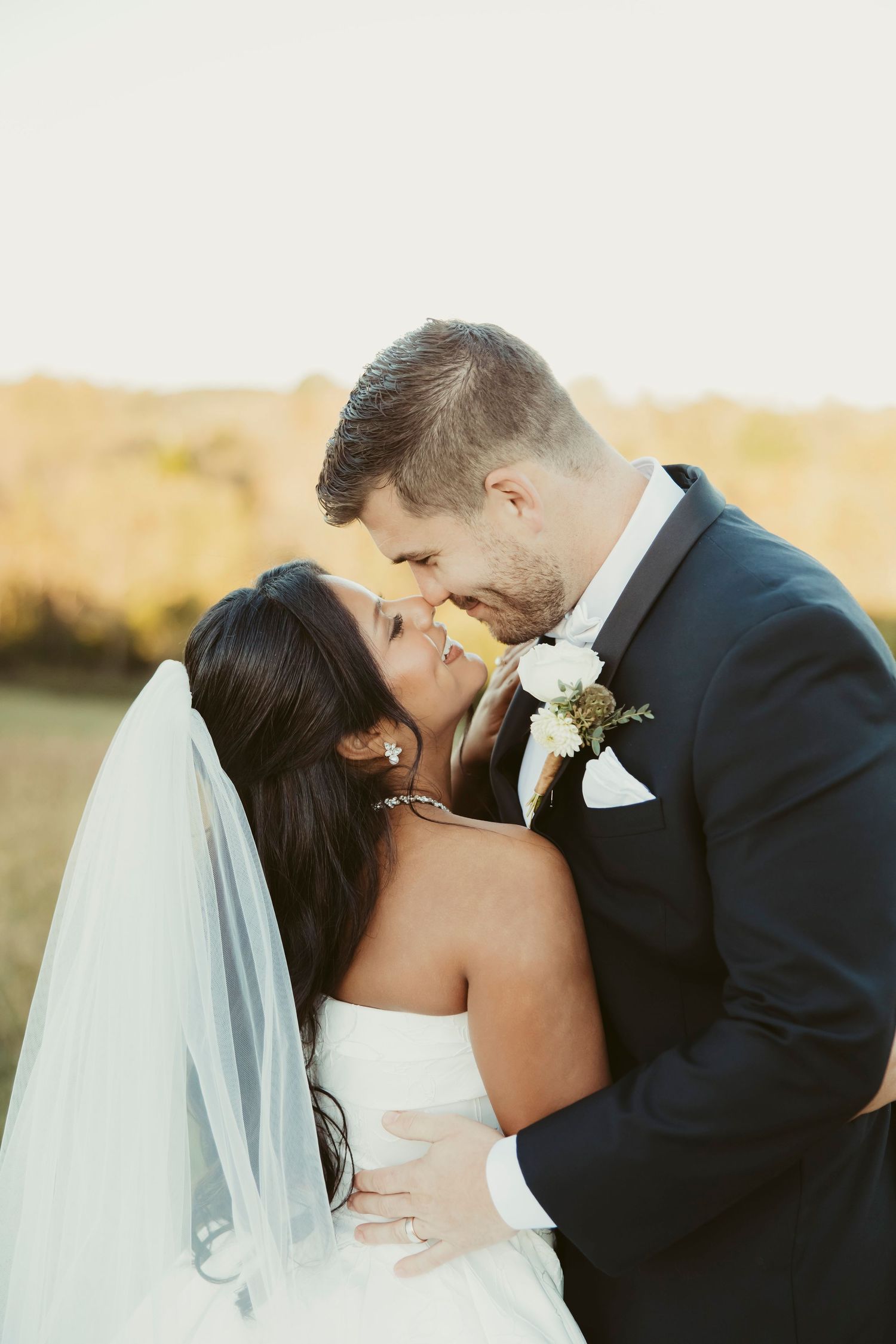 A bride in a white strapless gown and veil shares an intimate nose-to-nose moment with her groom in a navy suit in a golden outdoor field.