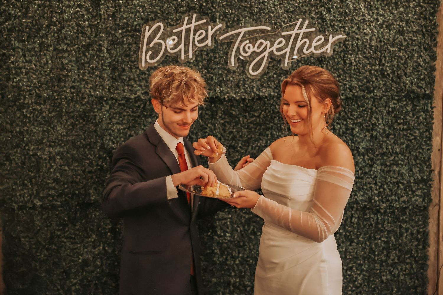 A couple in wedding attire dance beneath an illuminated 'Better Together' sign against a greenery backdrop.