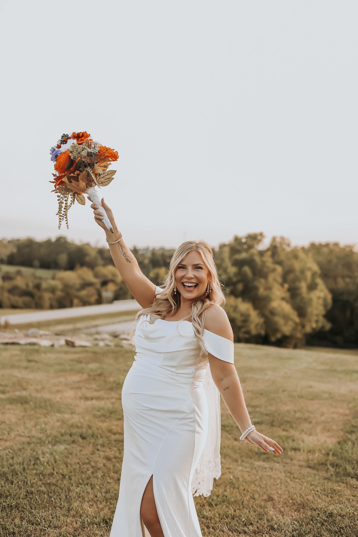 Joyful bride raises bouquet while celebrating outdoors in fitted white dress.