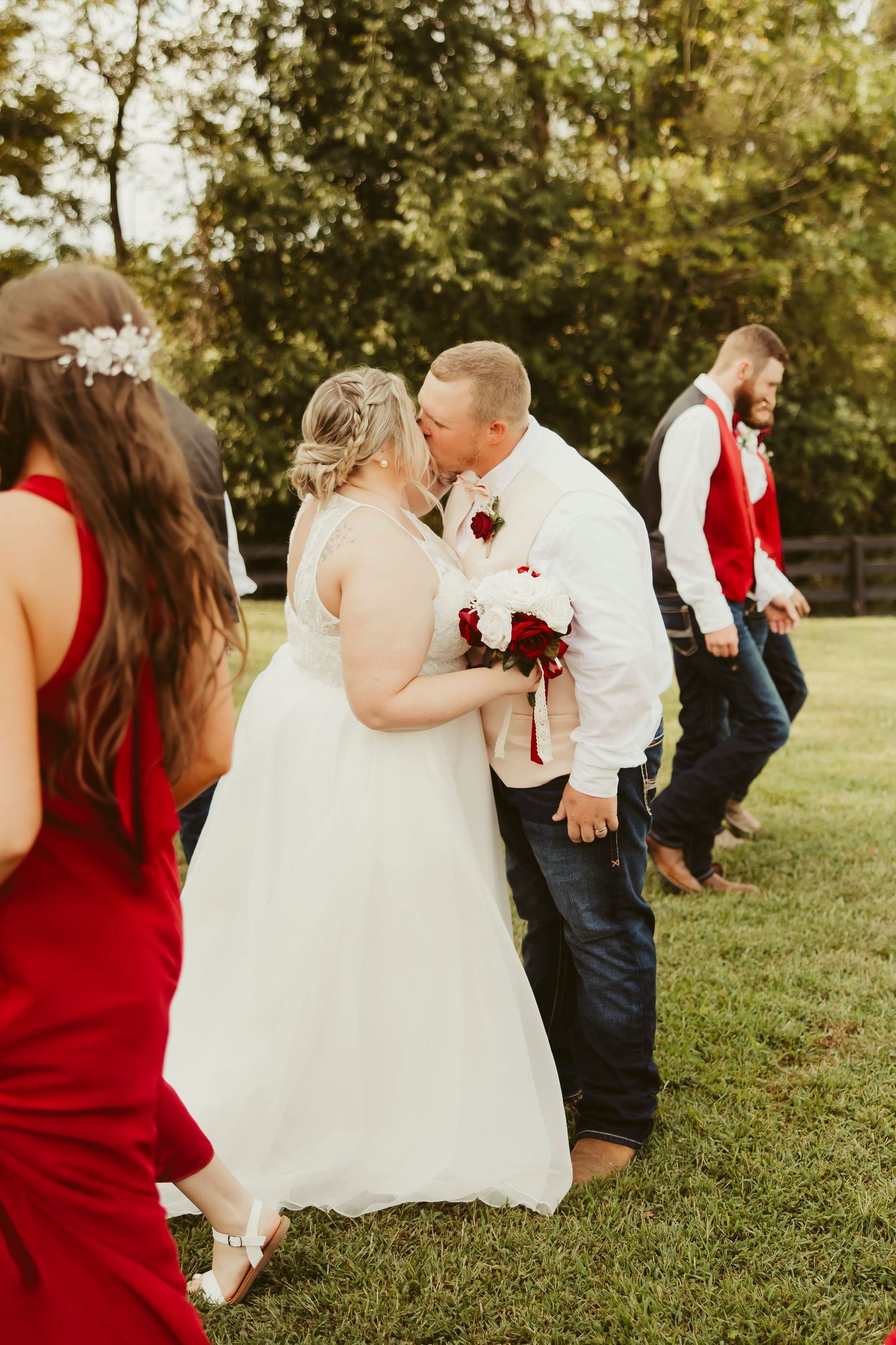 Bride and groom share a kiss outdoors on green grass, surrounded by wedding party in red dresses and vests.
