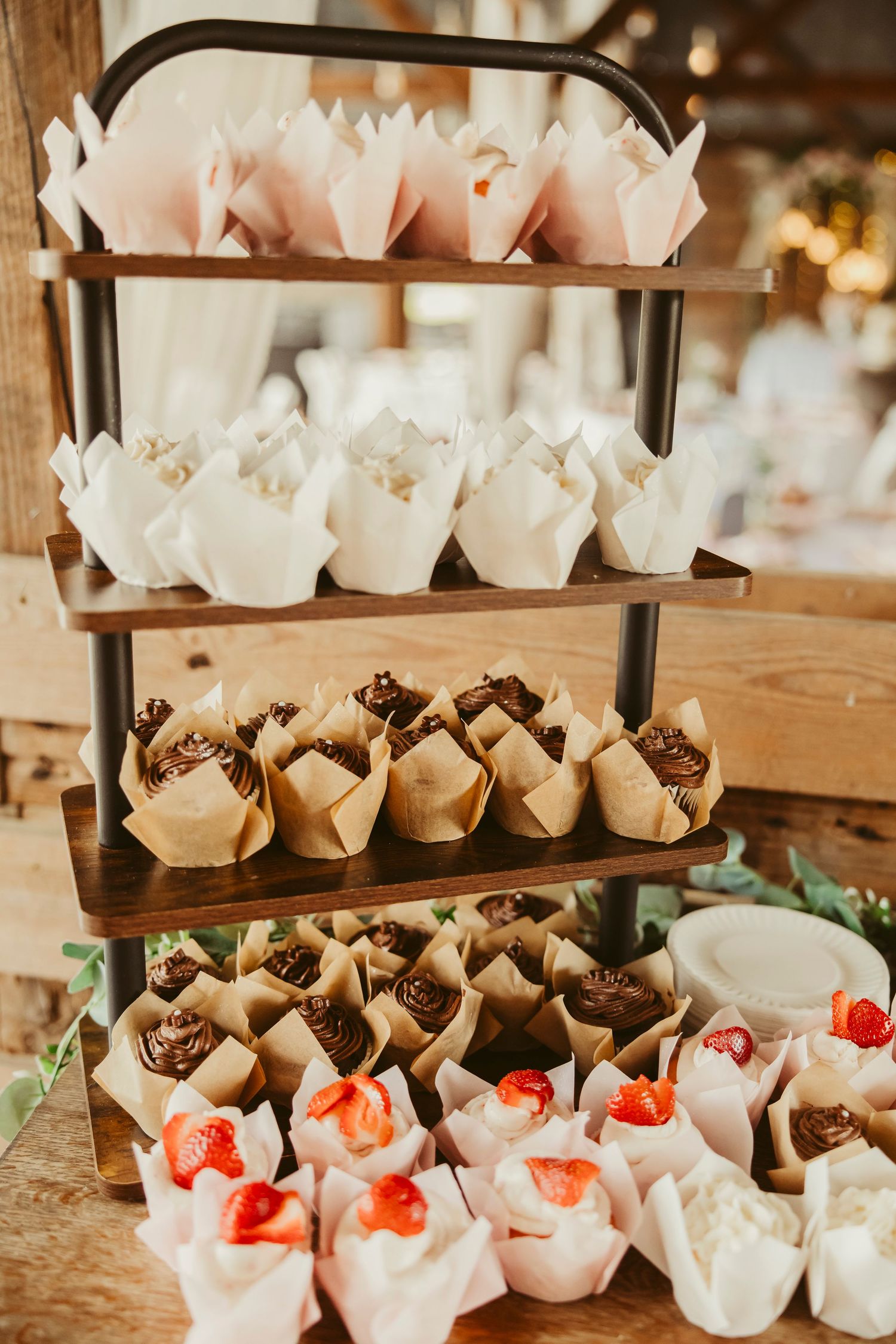 Three-tier wooden dessert stand displaying assorted cupcakes with strawberry and chocolate toppings in decorative paper wrappers.