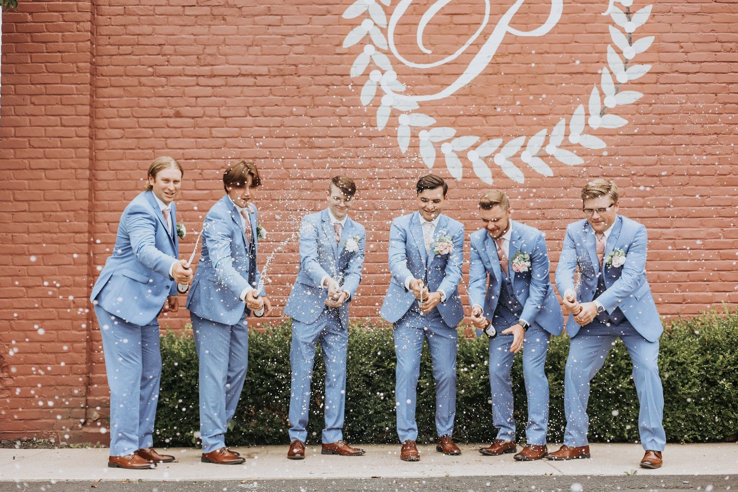 Groomsmen in light blue suits pose playfully against a brick wall with monogram decoration.