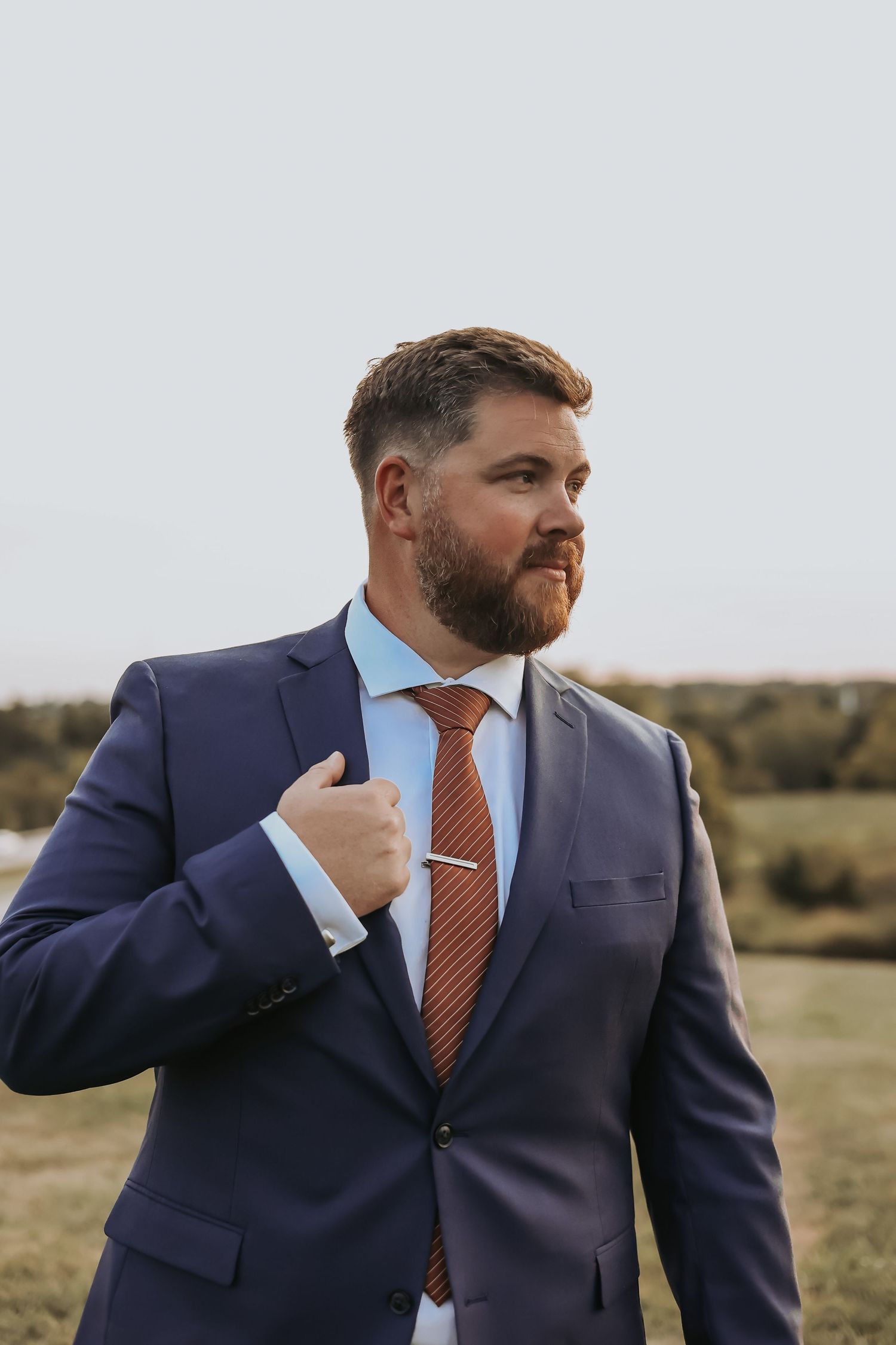 Groom in navy suit and orange tie poses against rural backdrop.