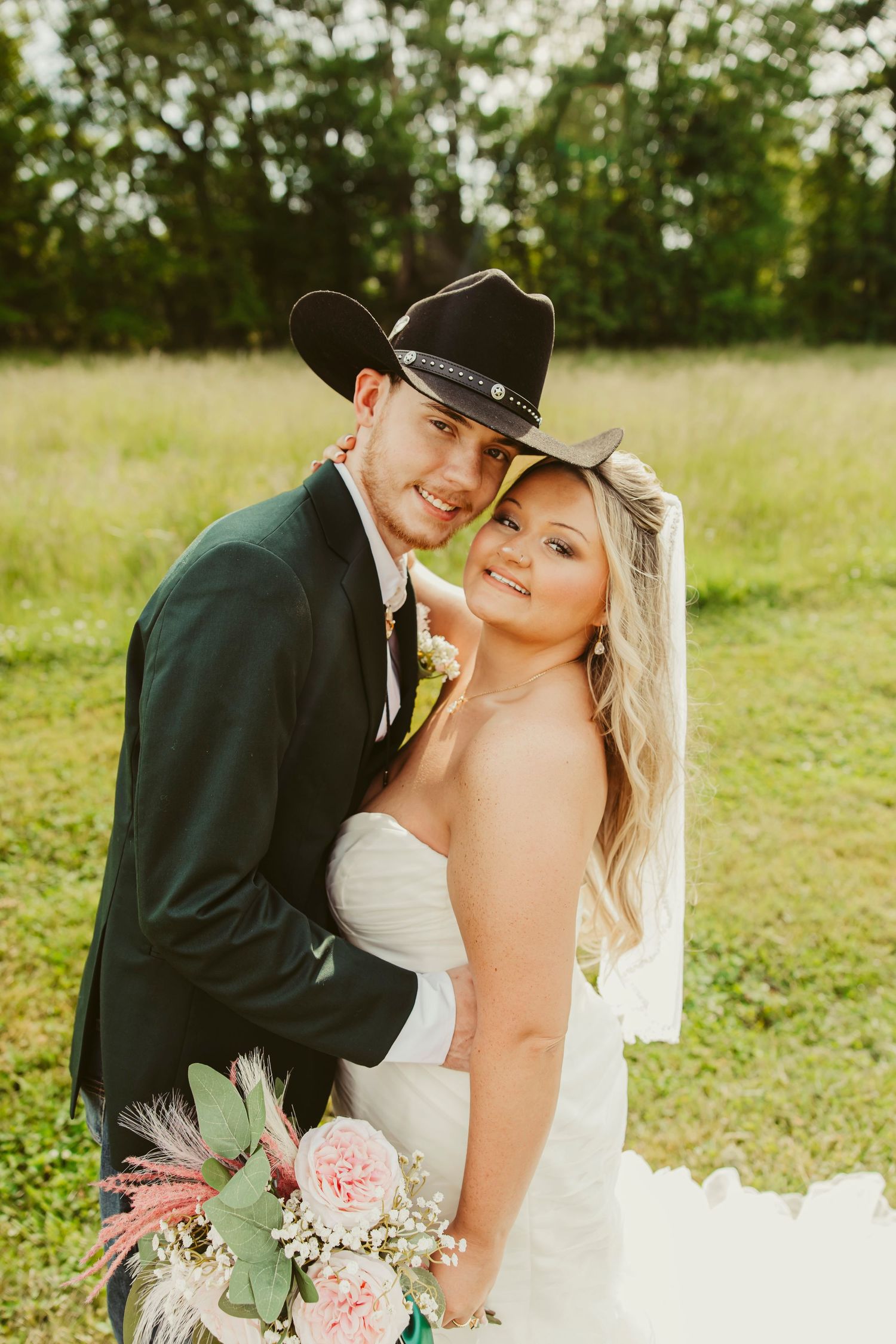 Bride and groom pose outdoors in a field, groom wearing a black cowboy hat, bride holding a pink floral bouquet.
