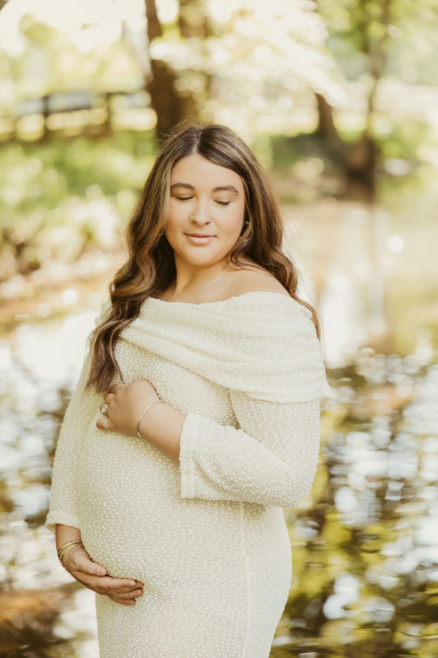 Pregnant woman in white off-shoulder dress cradling baby bump outdoors with soft bokeh green background.