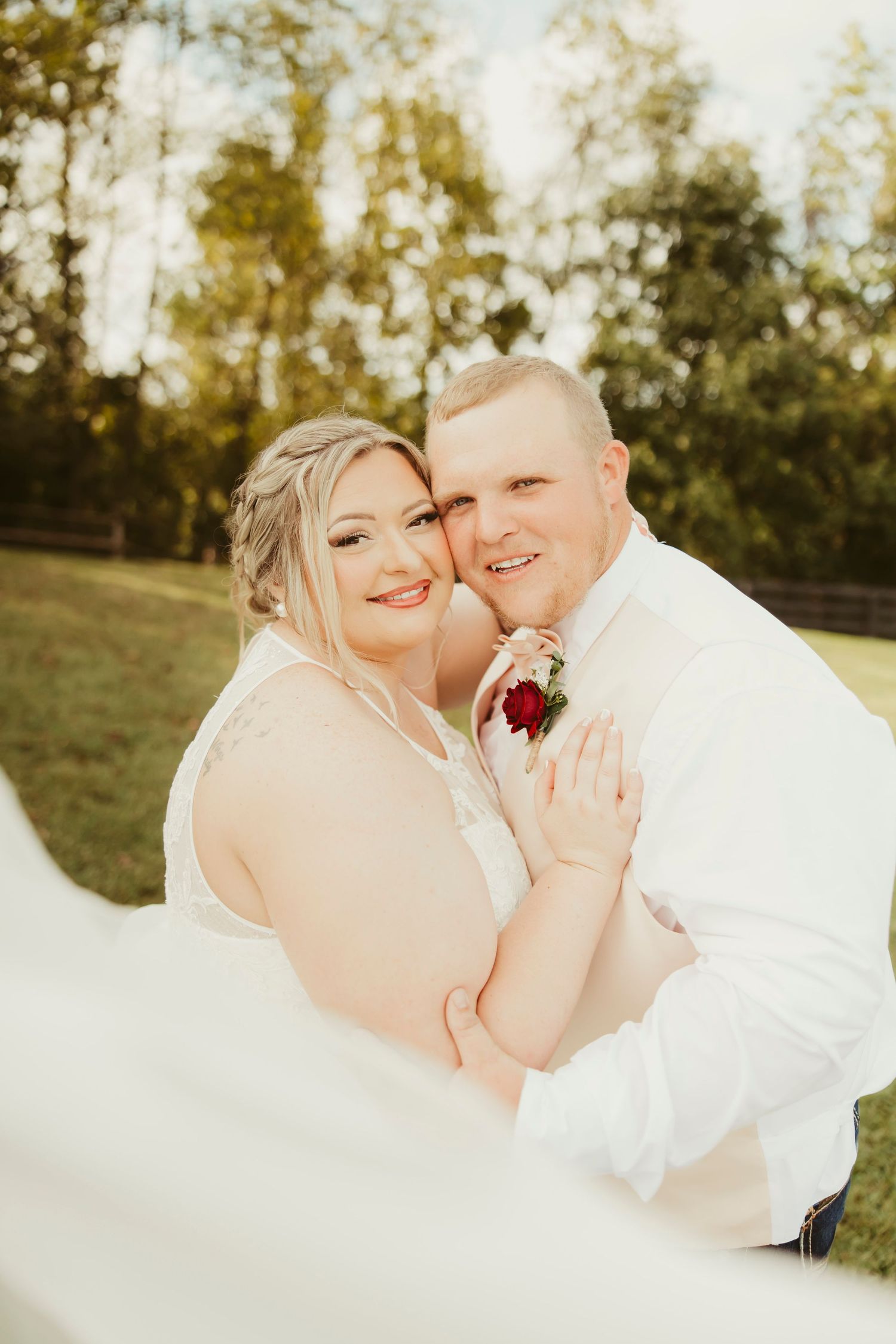 A smiling bride and groom embrace outdoors on their wedding day, with a flowing veil and red rose boutonniere.
