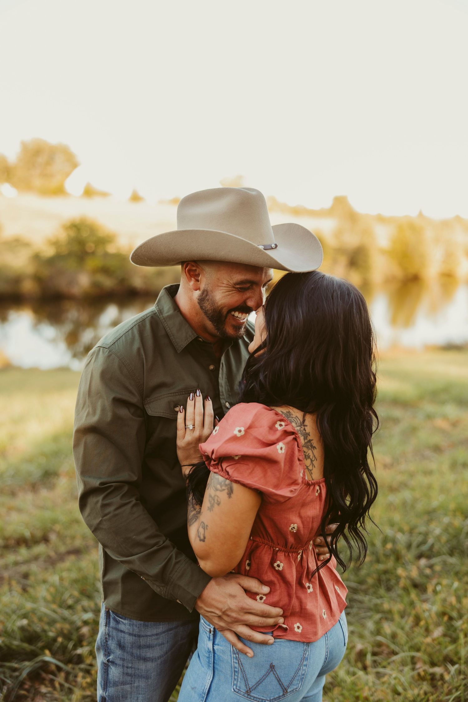 A smiling couple in cowboy hats share a romantic moment outdoors near a pond at golden hour.