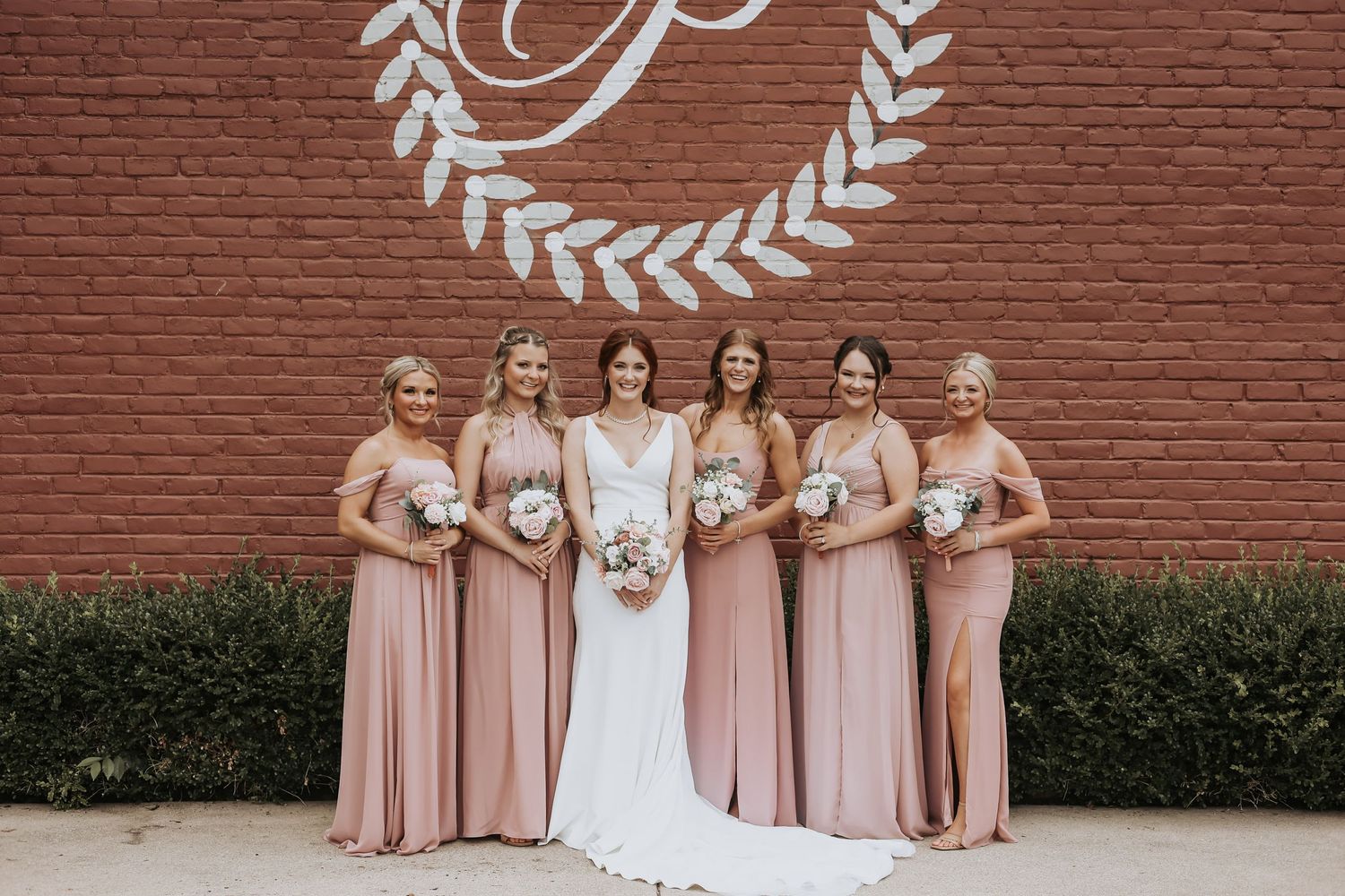 Bridesmaids in blush pink dresses pose with bride against brick wall with monogram.