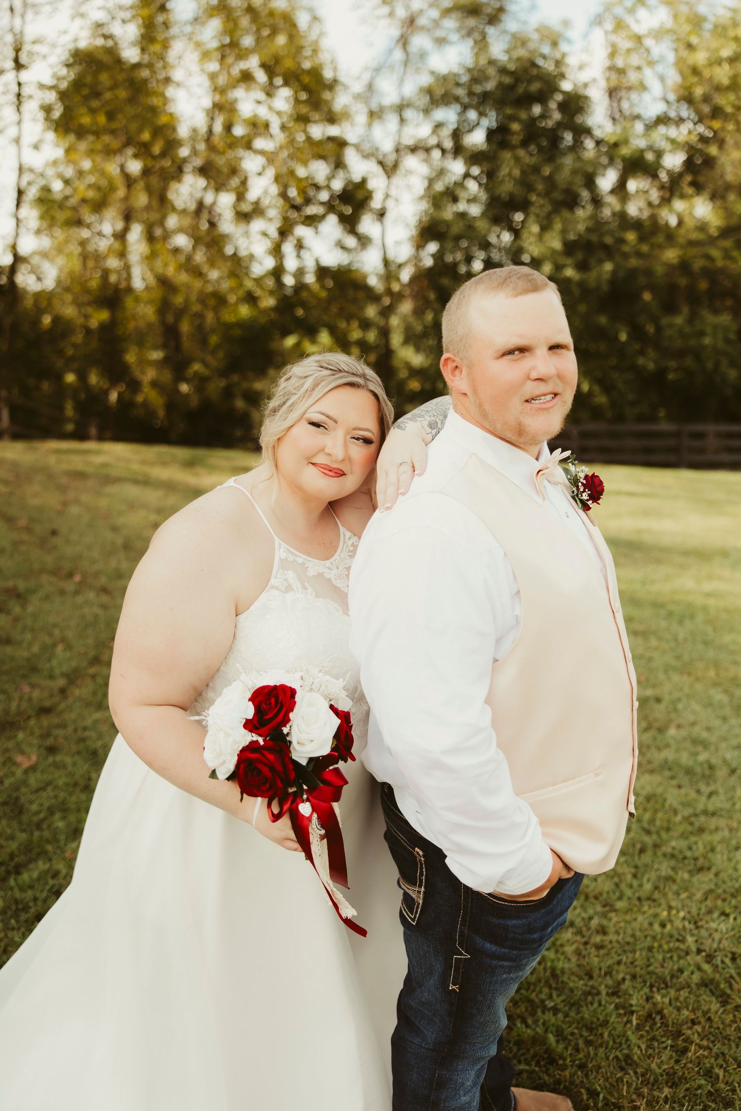 Bride and groom pose outdoors in golden light, she holds red and white rose bouquet, he wears white shirt.