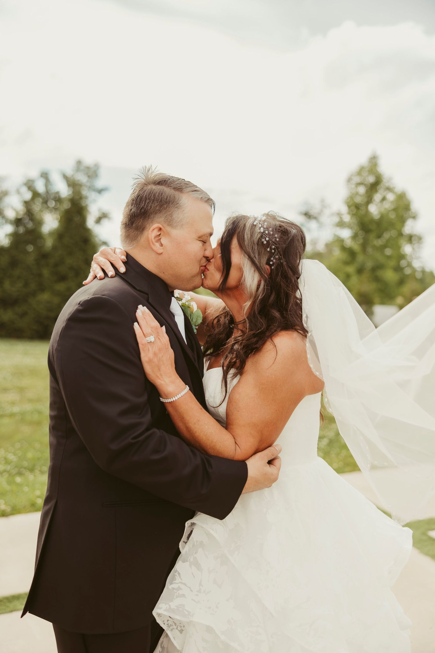 Bride and groom share a passionate kiss outdoors, with the bride's veil flowing in the wind.